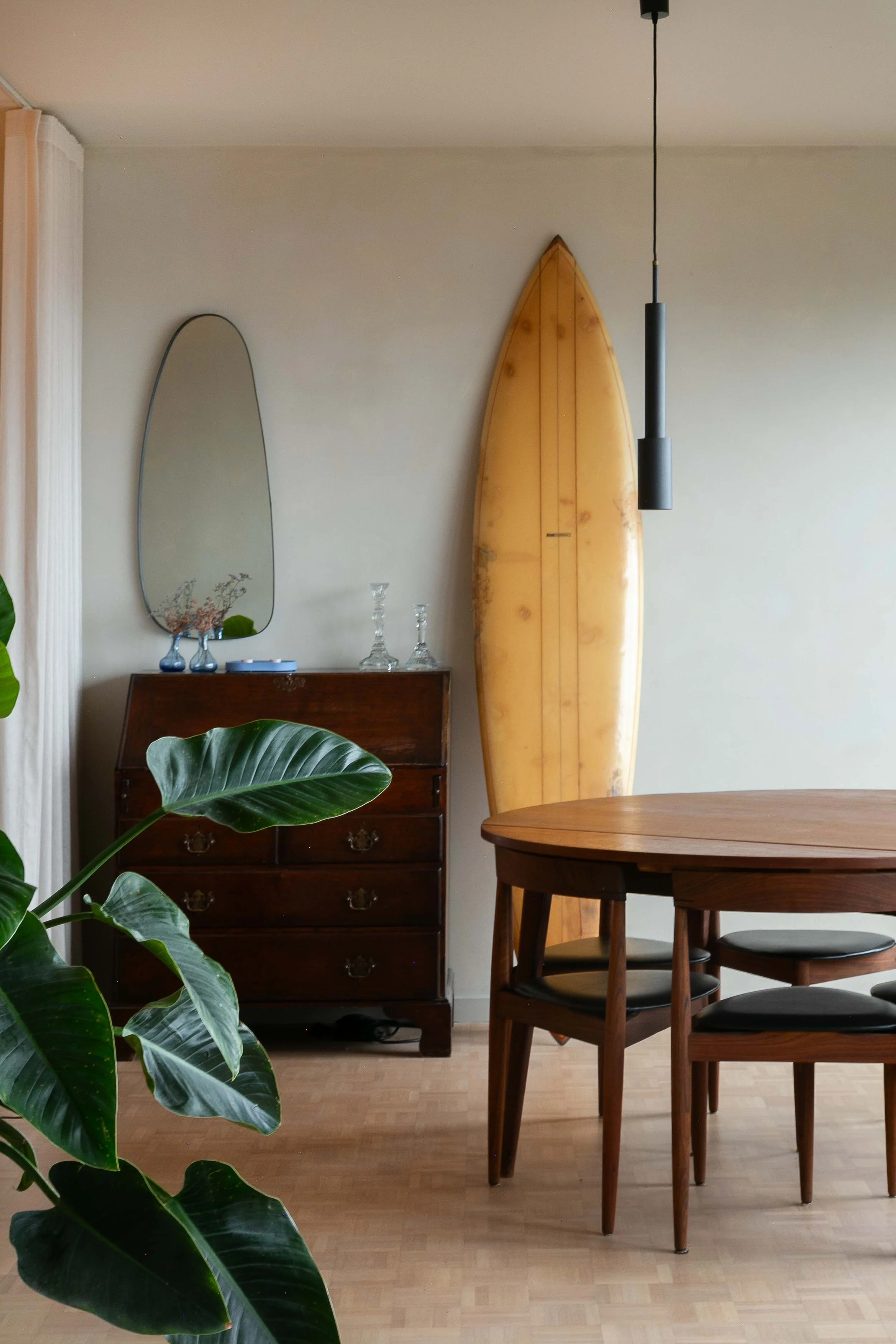 A dining area with a wooden table and four chairs, a vintage dark wooden dresser with decorative glassware on top, a tall oval mirror, a surfboard leaning against the wall, a black pendant light hanging from the ceiling, a leafy green plant in the fo