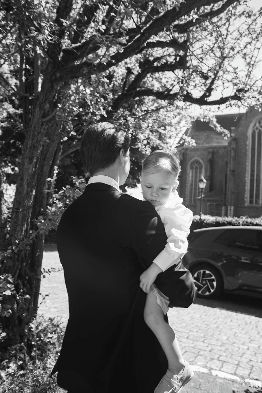 A man in a suit holding a young boy outdoors near a tree and a church, with parked cars in the background.