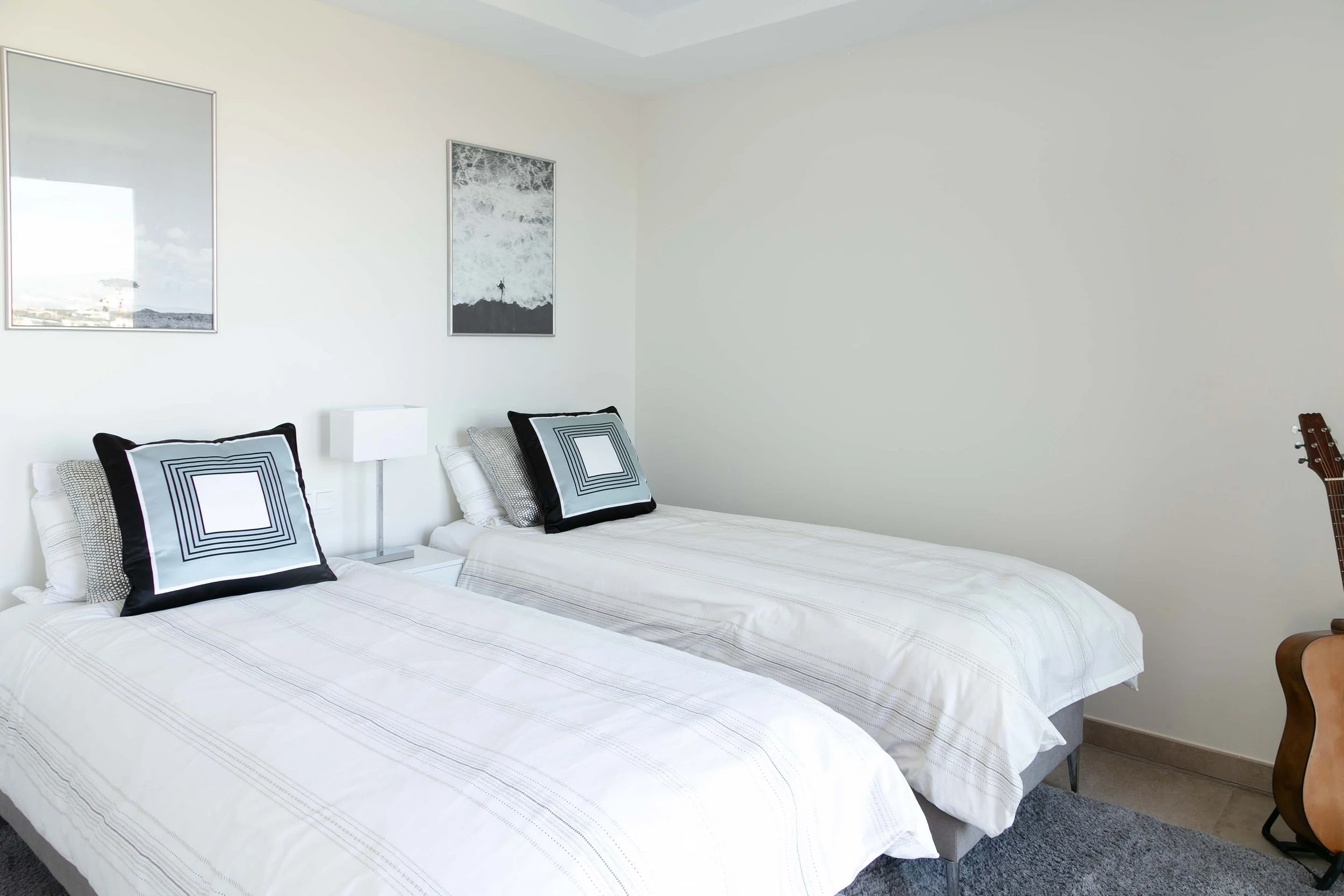 Two single beds with matching white and gray striped bedding and decorative pillows, a nightstand with a lamp, framed artwork on the white wall, a guitar resting against the wall, and a small part of a chair.