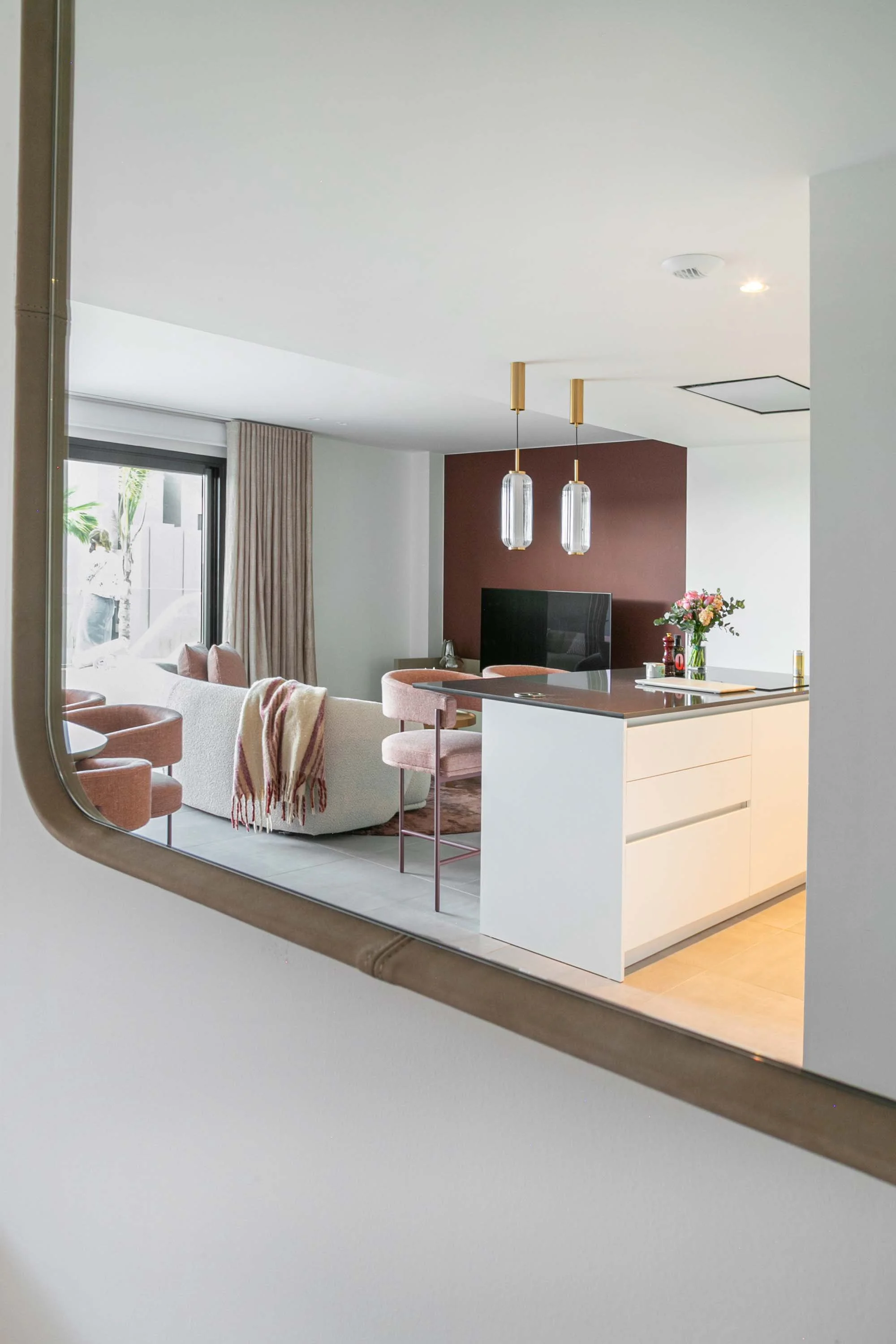 A modern open-concept living space viewed through a rounded rectangular mirror, showing a cozy seating area with pink chairs and a white sofa, a kitchen island with a flower arrangement, and a dark accent wall with a television.