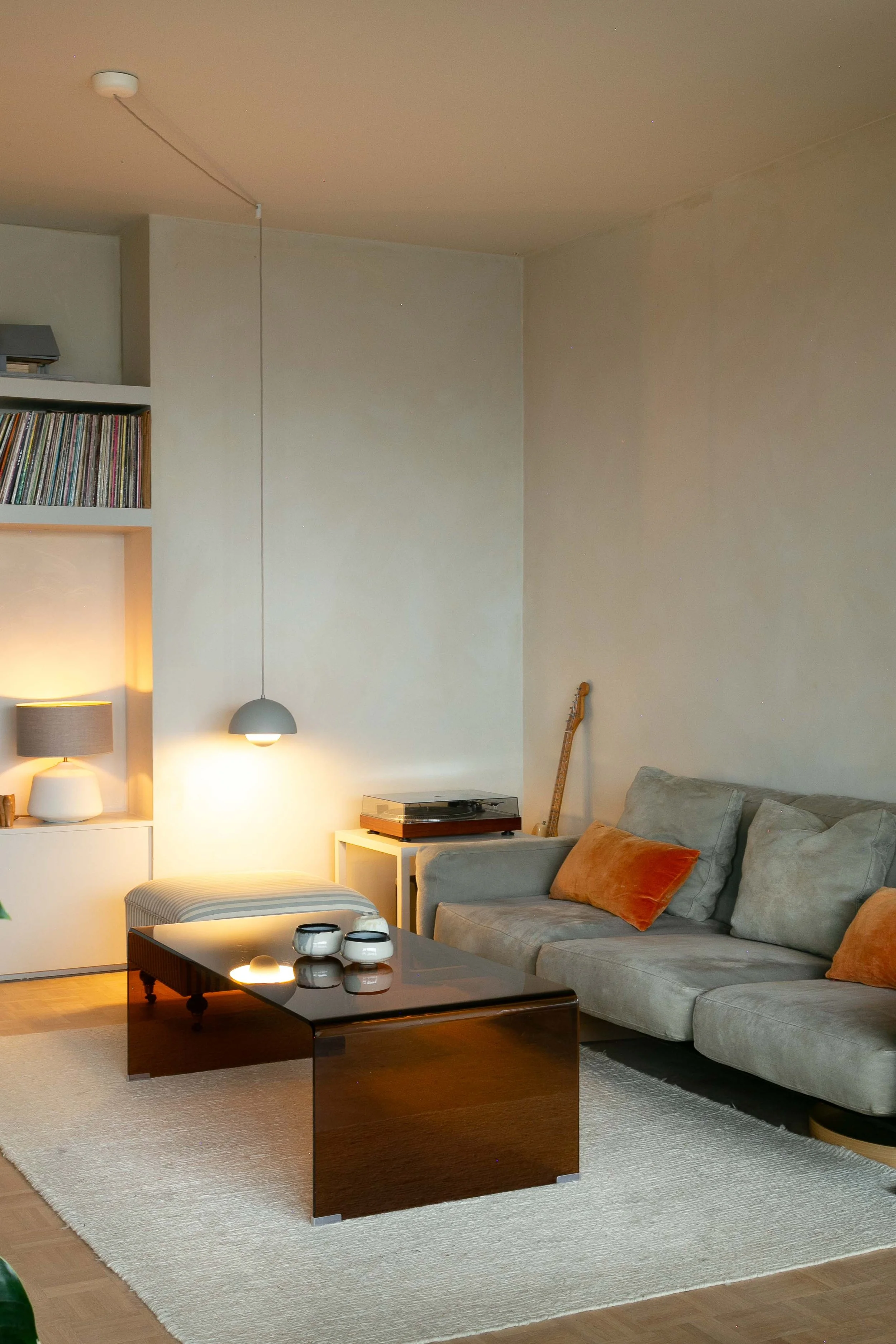 Modern living room with a beige sofa, orange and beige cushions, a black coffee table, a white side table with a turntable, a lamp on the side table, a bookshelf with vinyl records, a floor lamp, a guitar in the corner, a hanging light, and a beige r