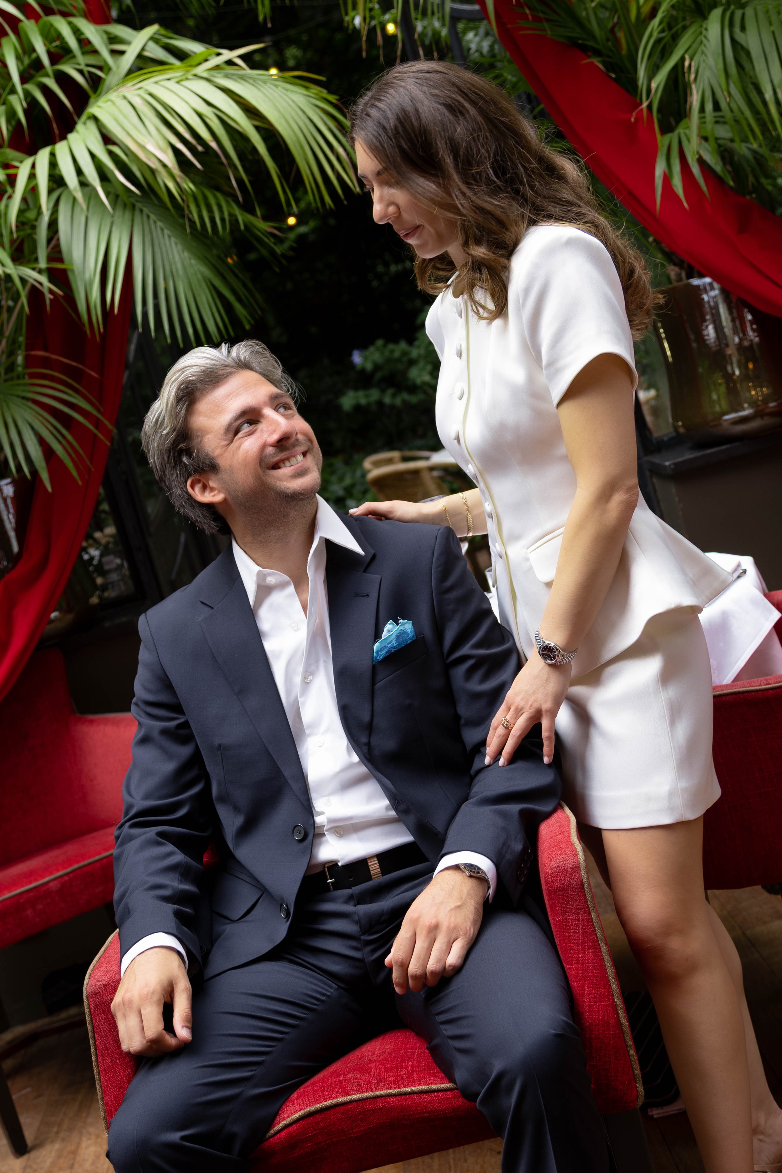 A man in a suit sitting on a red velvet chair and smiling at a woman standing next to him, indoors with green plants and red fabric drapery in the background.
