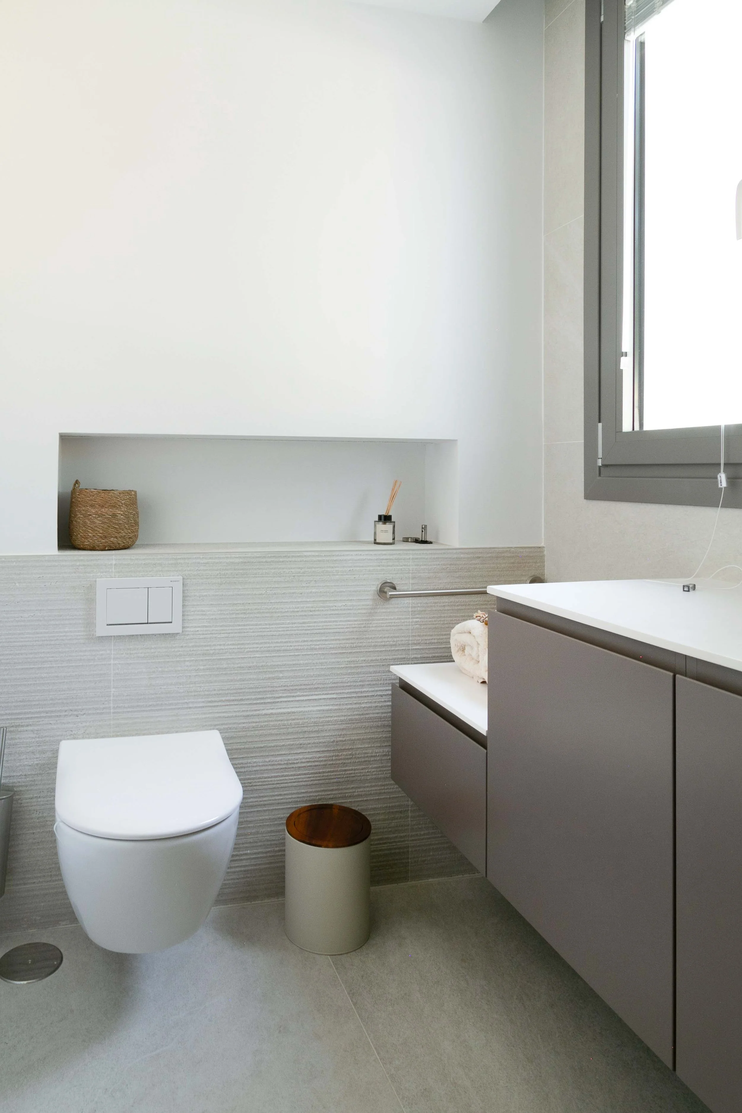 Modern bathroom with gray cabinets, a white toilet, a small round trash bin with a wooden top, a recessed shelf with a basket and toiletries, a window with gray trim, and a textured wall behind the toilet.