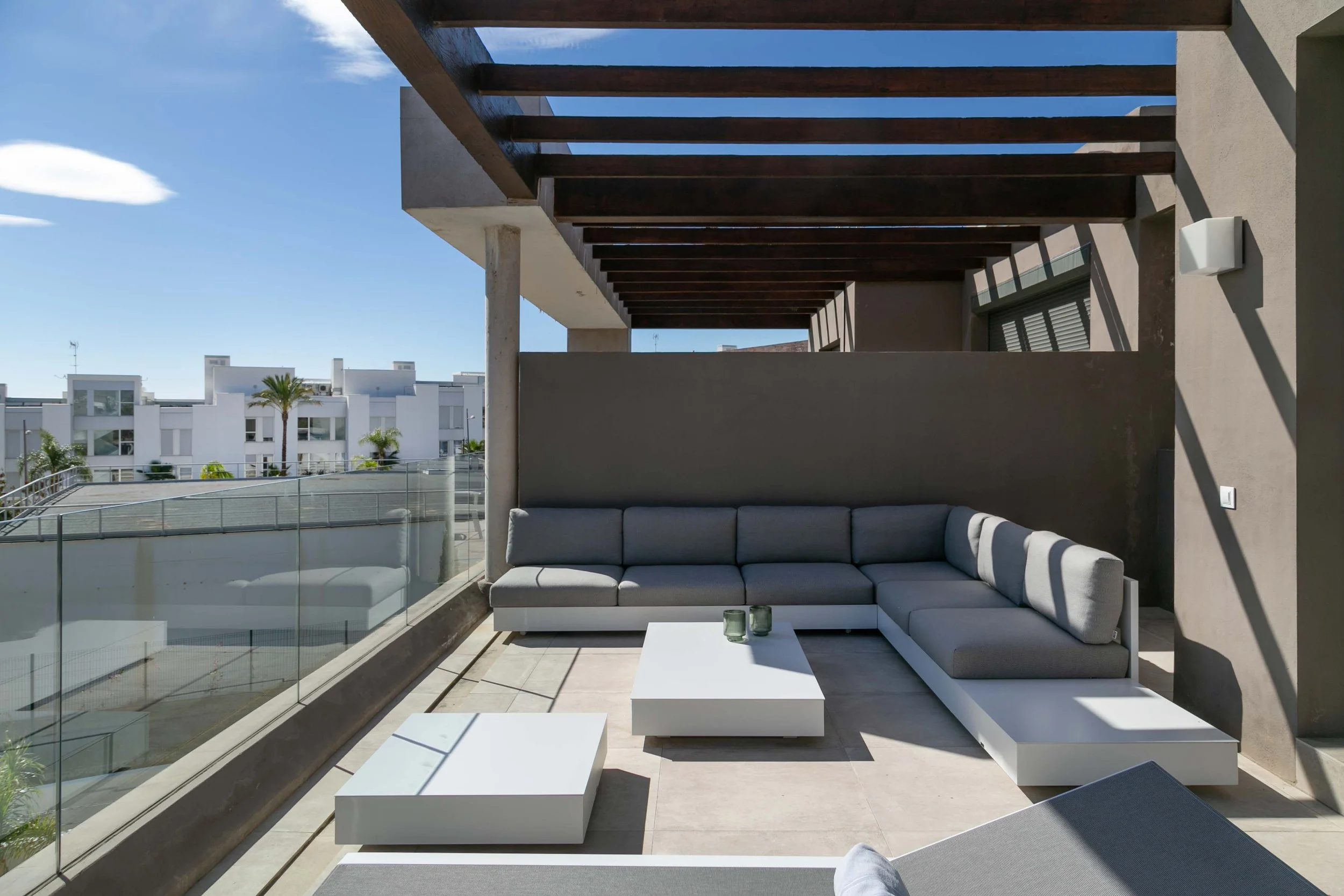 Modern outdoor balcony with gray sectional sofa, white coffee tables, and a glass railing. Overhead wooden beams and a view of white buildings and palm trees under a sunny sky.