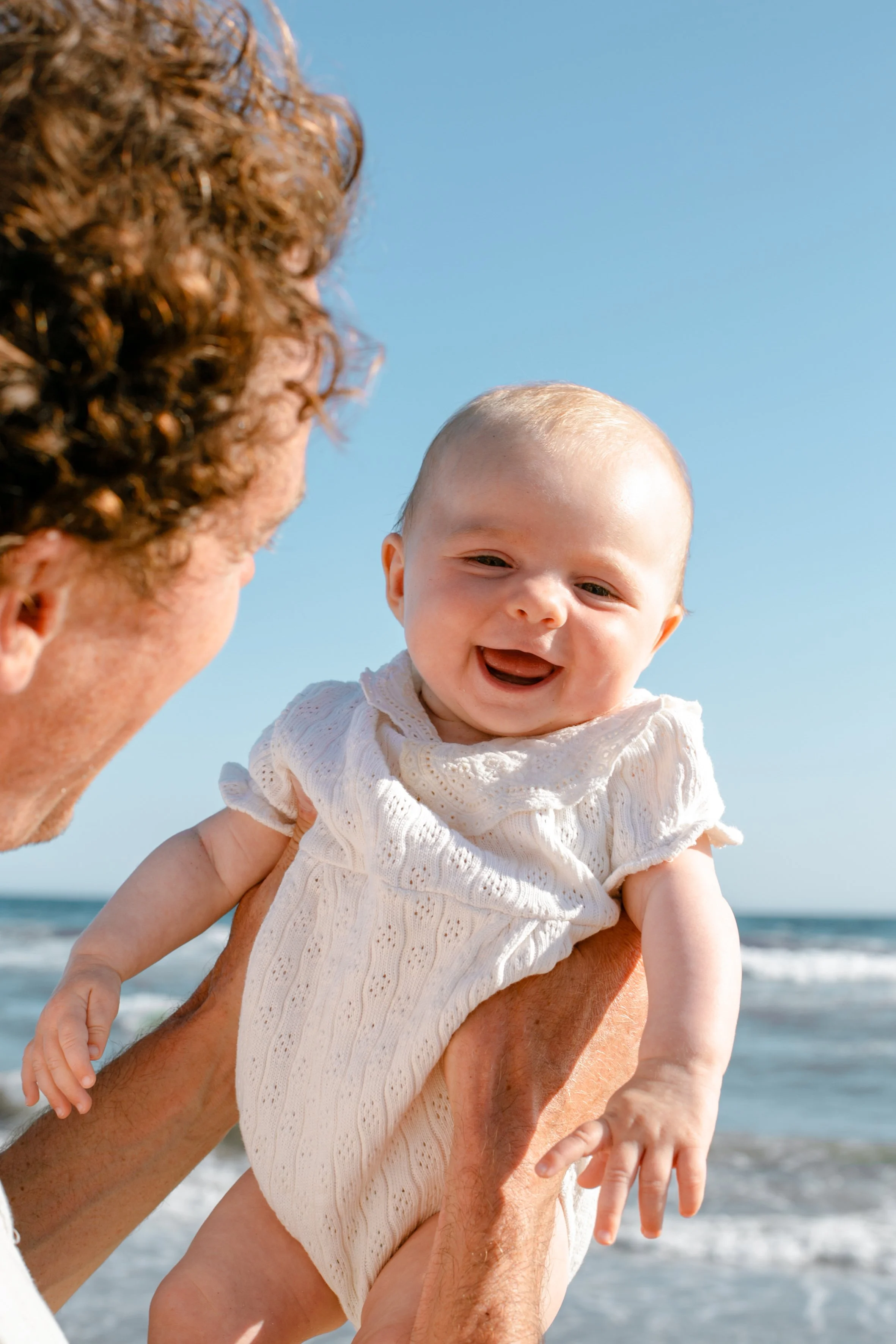 A man holding a smiling baby girl at the beach with ocean waves and blue sky in the background.