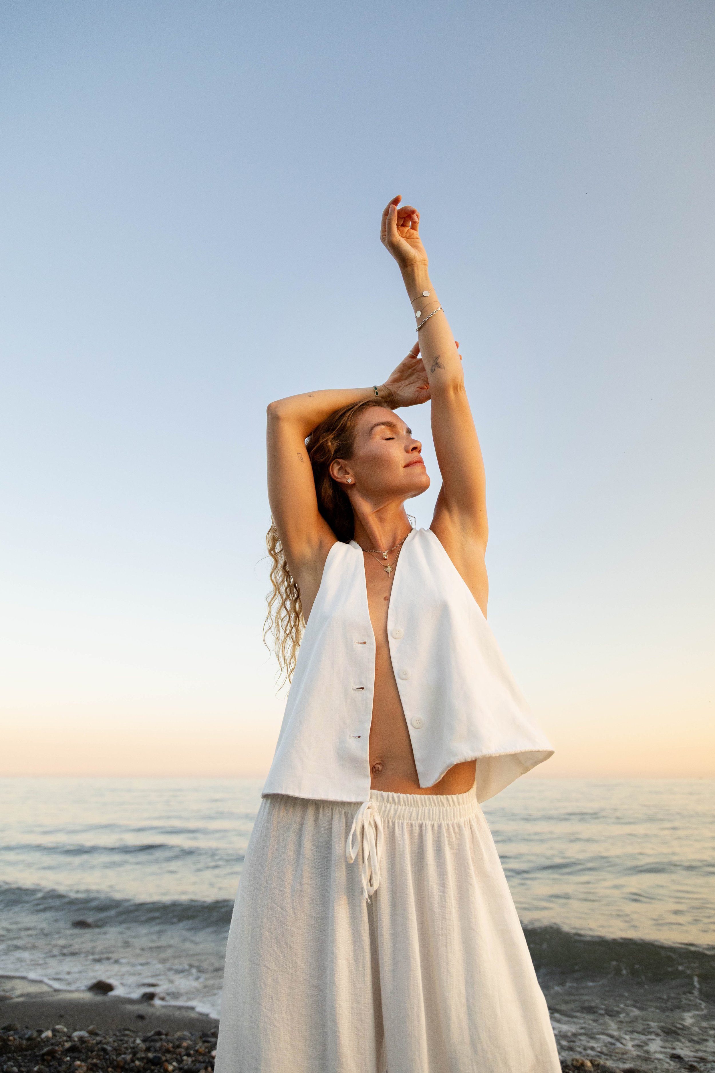 A woman with closed eyes, wearing a white sleeveless top and cream-colored wide-leg pants, stands on the beach with her arms raised above her head in a relaxed yoga pose during sunset or sunrise, with the ocean and sky in the background.
