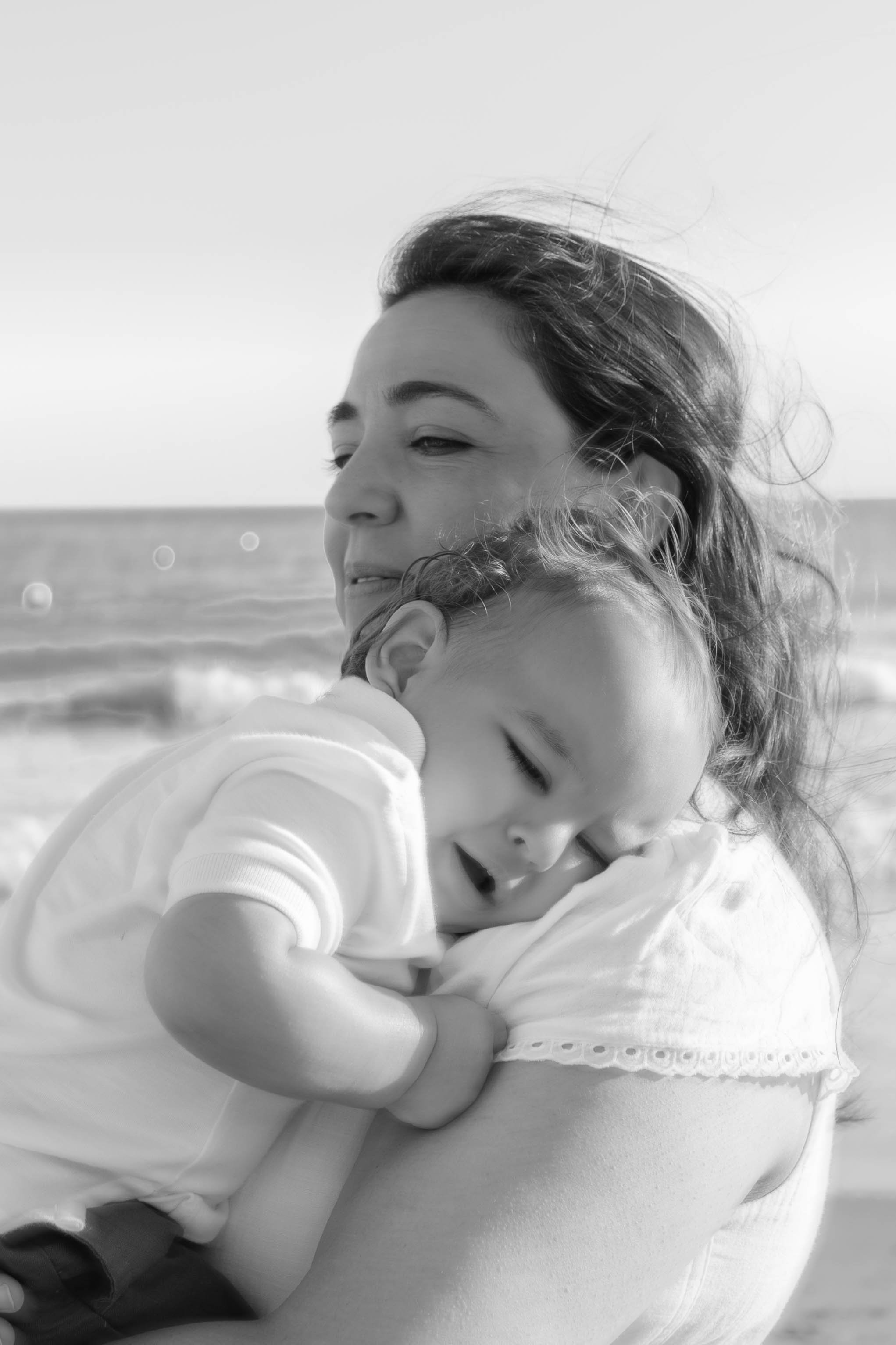 A woman and a young girl are embracing each other at the beach, with the woman gently holding the girl and both with closed eyes, conveying a moment of affection and tenderness.