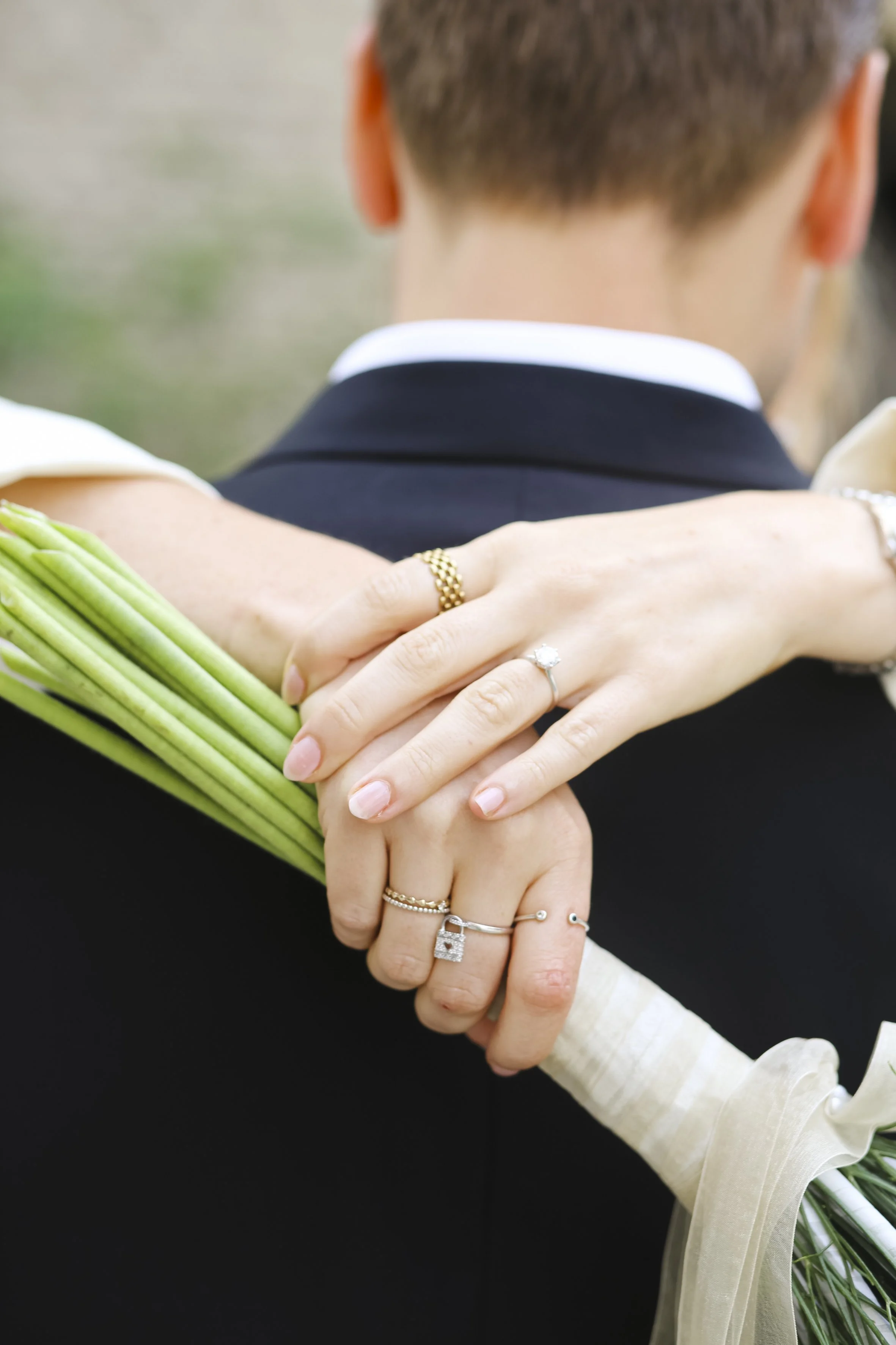 Close-up of a person wearing rings, holding a bouquet of green flowers, with someone else embracing them in an outdoor setting.