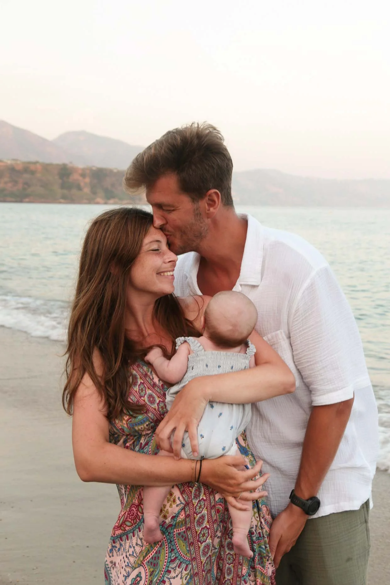 A family of three on the beach, with a woman holding a baby, a man kissing her forehead, and a scenic water and mountain background. There first beach photography session in marbella