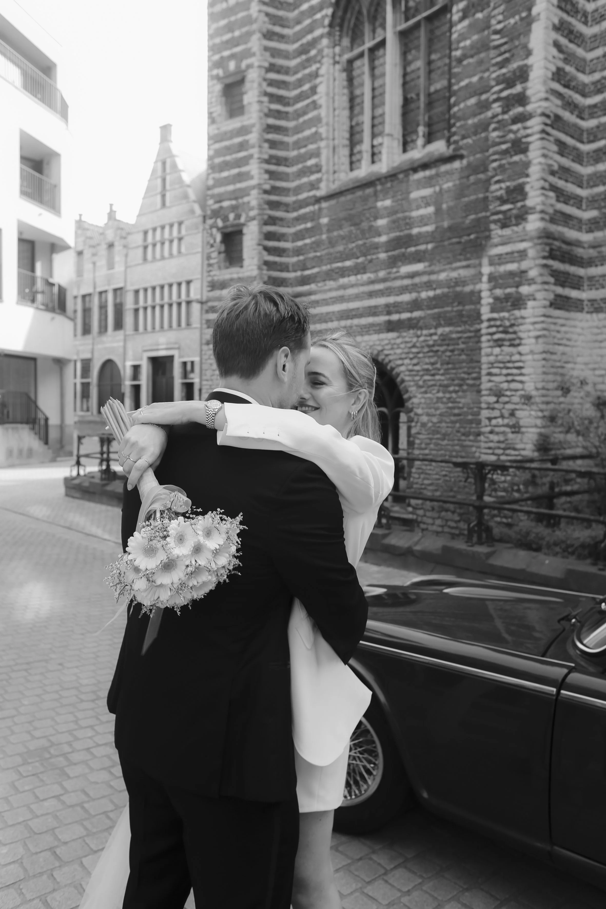 A couple in wedding attire hugging outdoors with a historic brick building in the background. The bride carries a bouquet of flowers.