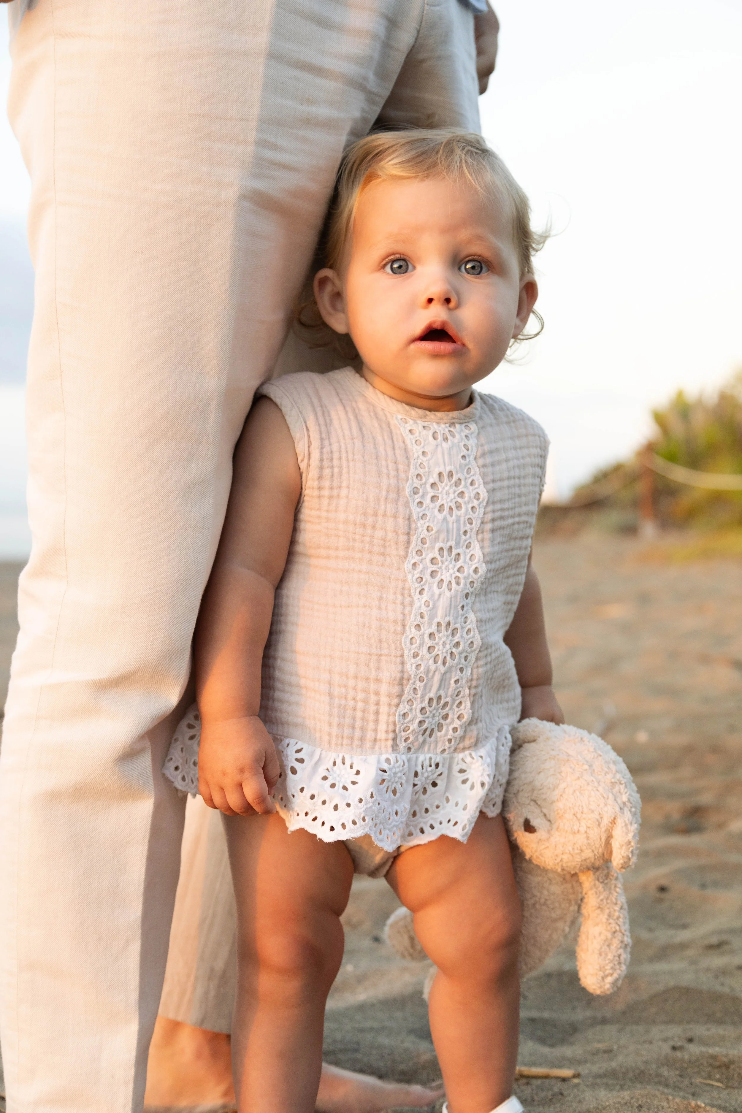 A young girl holding a teddy bear on a beach at sunset, with an adult partially visible next to her.