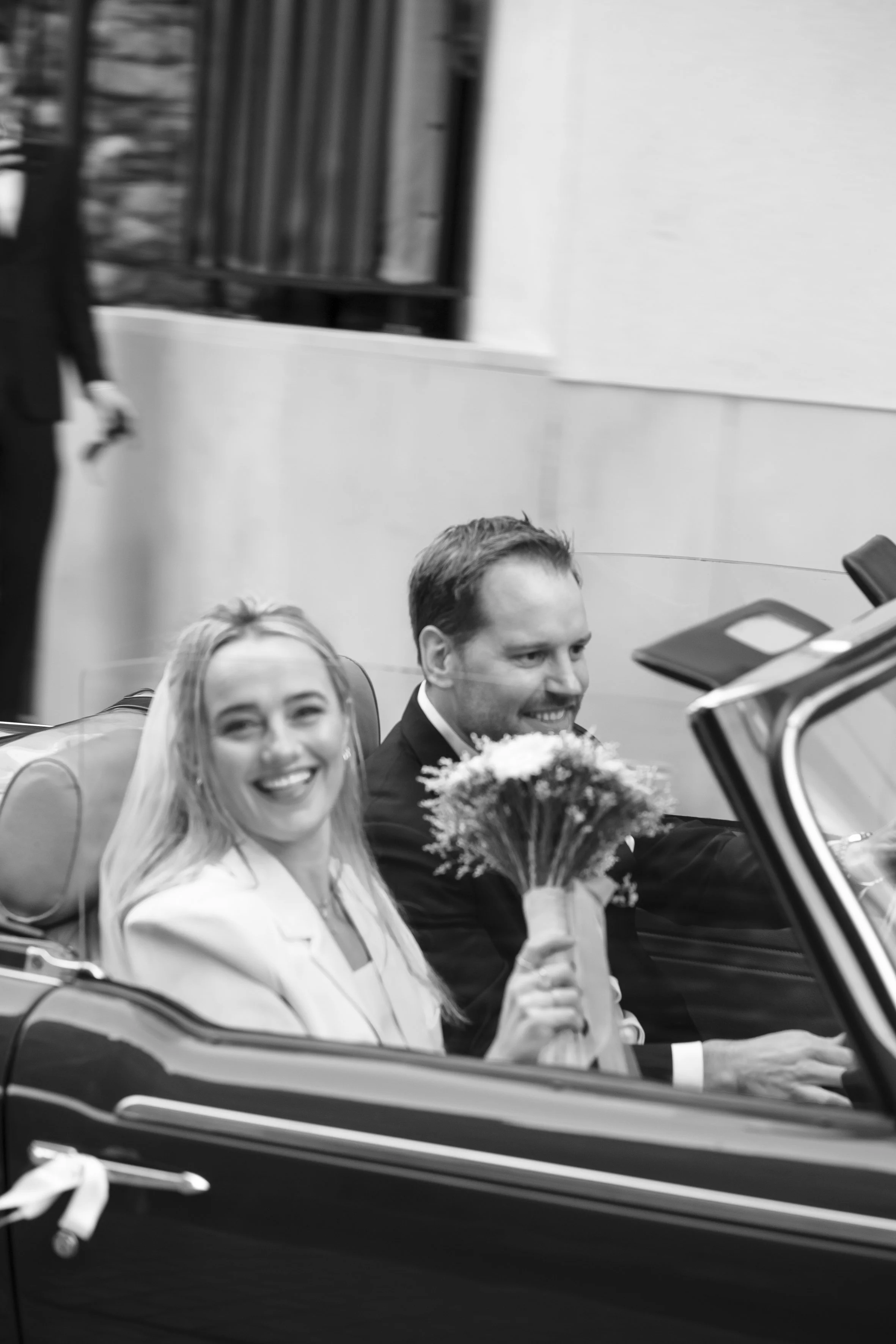 A black-and-white photo of a smiling bride and groom in a convertible car, with the bride holding a bouquet of flowers. They appear to be at a wedding celebration.