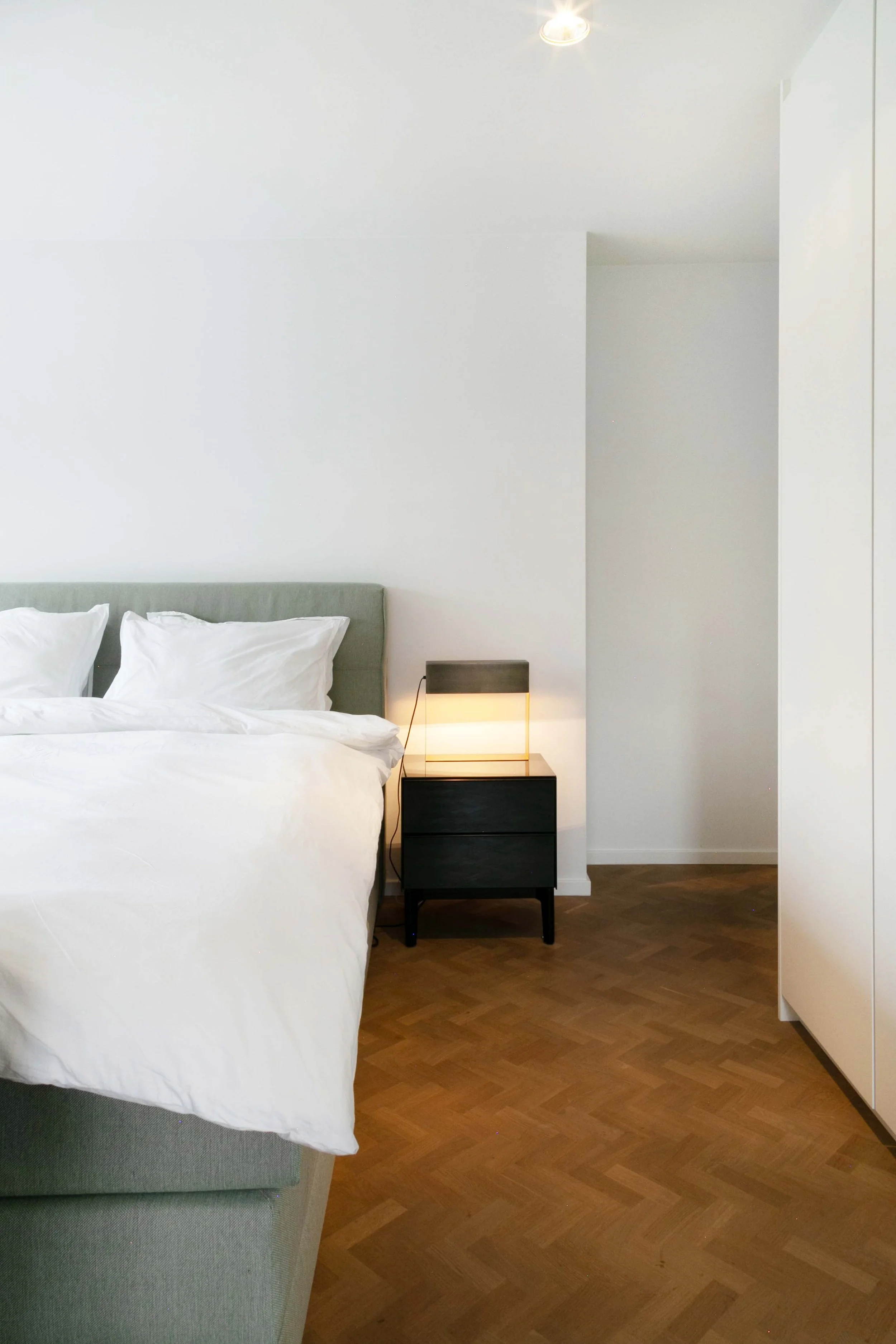 A minimalist bedroom with white walls, a bed with white bedding, a gray upholstered headboard, a small black nightstand with a modern lamp, and hardwood flooring.