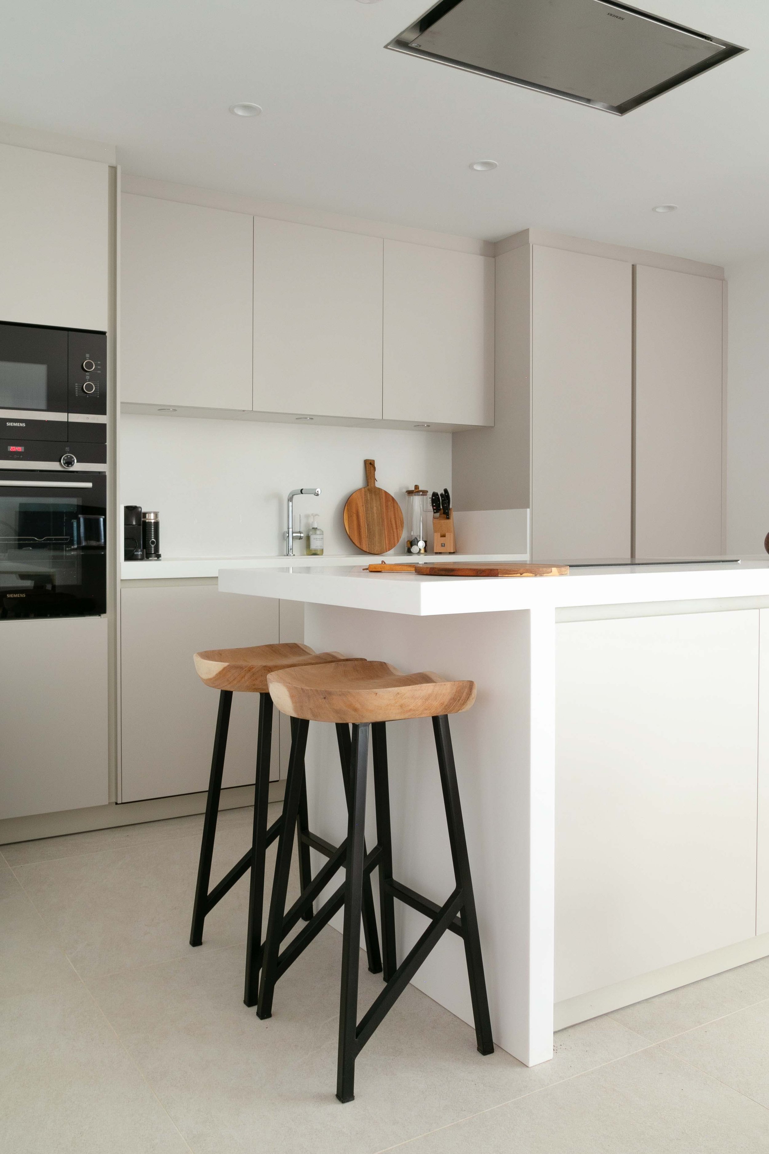 Modern kitchen with white cabinets, black stools, a white countertop, and built-in appliances. There are wooden cutting boards, knives, and small kitchen items on the counter.