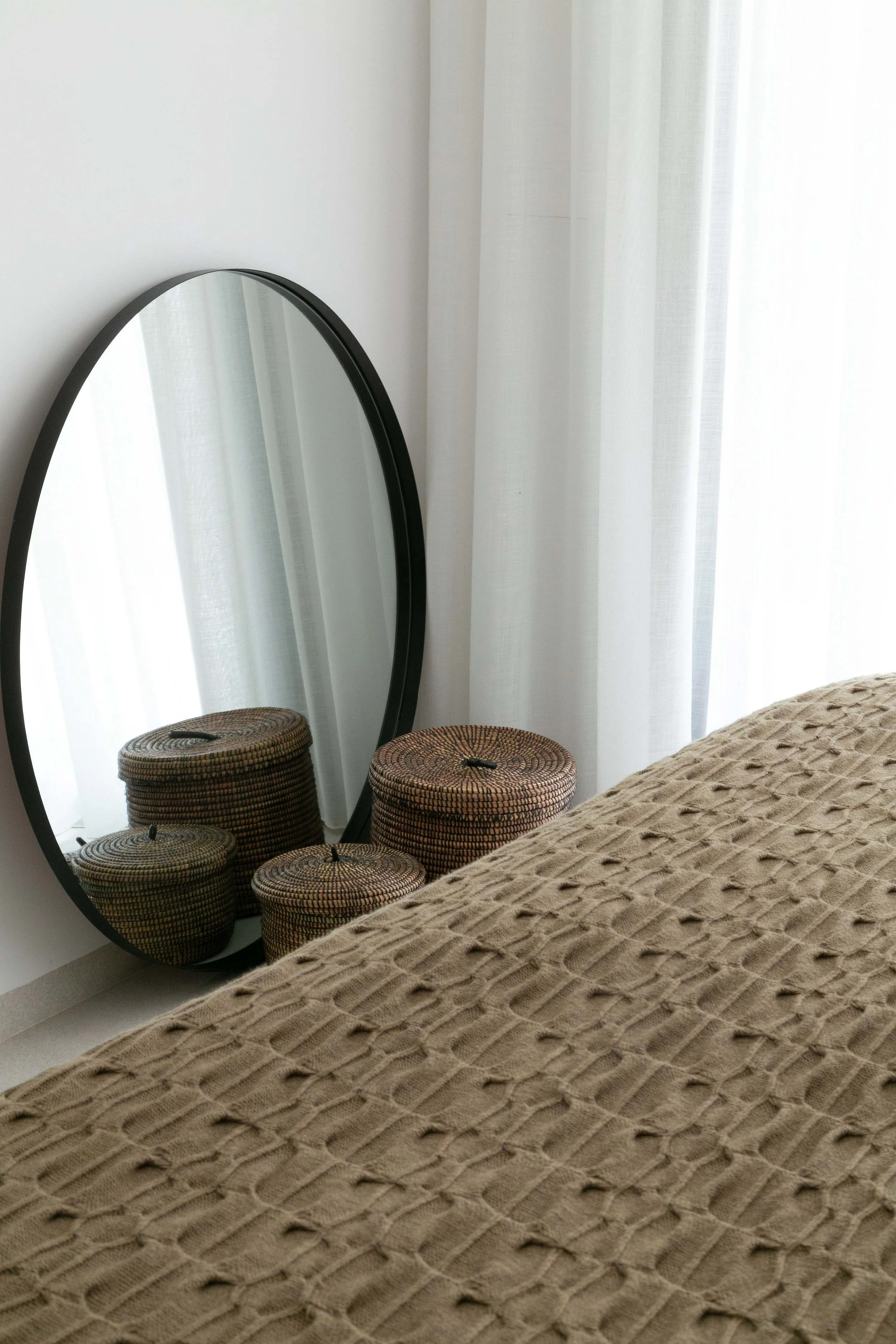 A bedroom corner with a standing mirror, woven baskets stacked below it, a beige textured blanket on a bed, and sheer white curtains blocking sunlight.