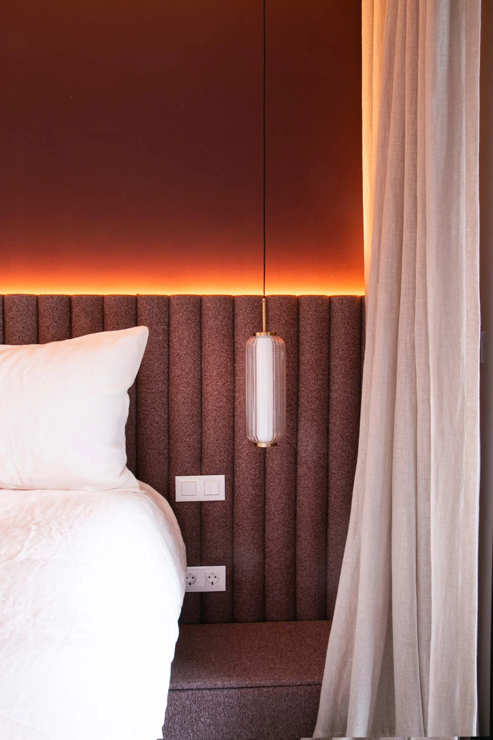 A hotel bedroom with a bed featuring white bedding, brown upholstered headboard, and a hanging pendant light on the wall. There are curtains on the right side and multiple electrical outlets on the wall.