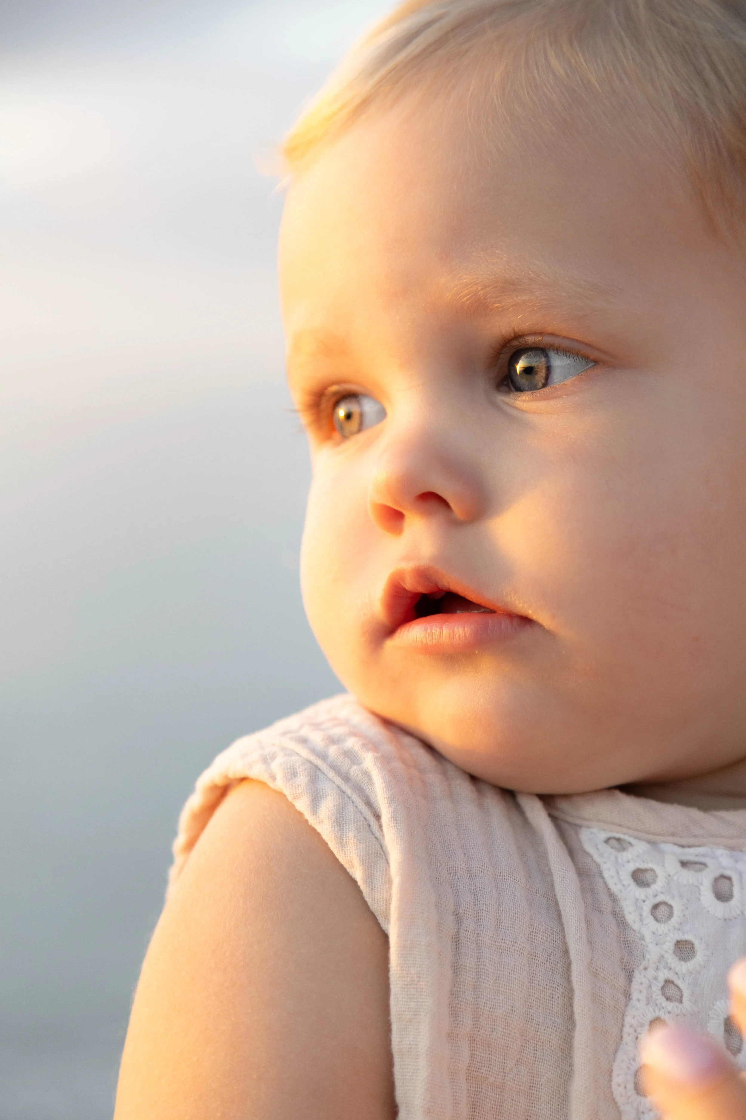 Close-up of a child's face and shoulder, with blonde hair and blue eyes, wearing a light-colored dress with eyelet lace details, illuminated by warm sunlight.