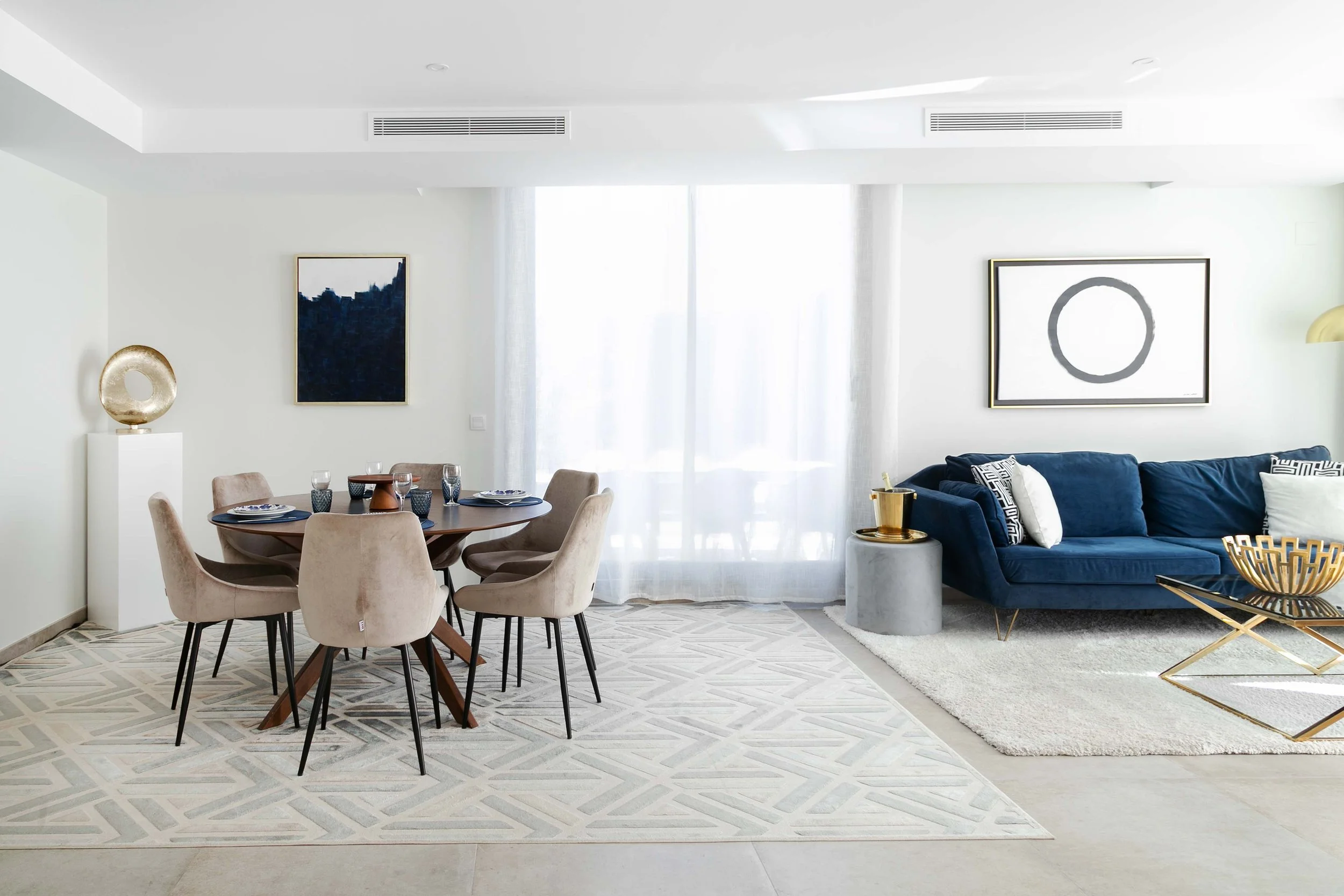 Modern living room with a dining area, beige chairs, a round table with plates and glasses, a blue velvet sofa, framed abstract art, a white wall, a gold decorative sculpture, and curtains letting light in.
