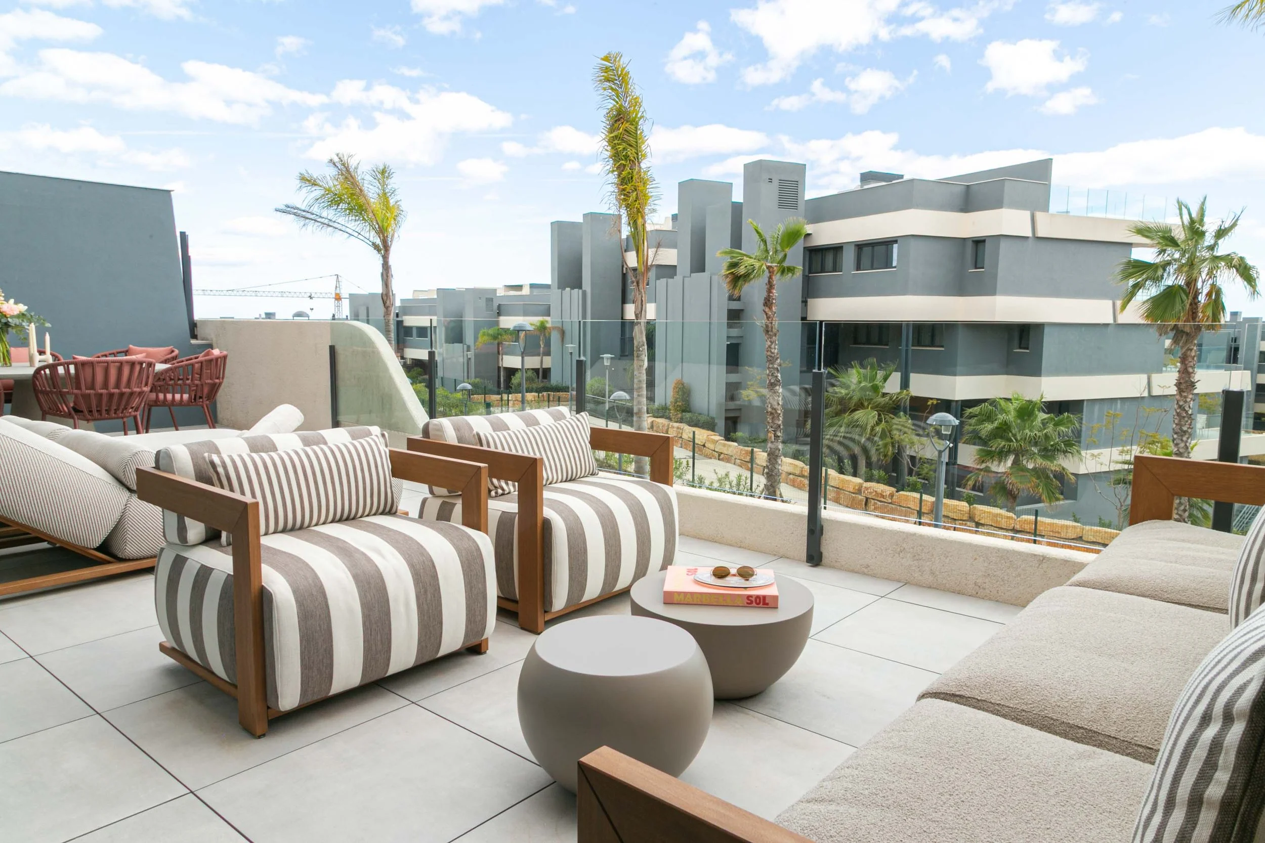 Balcony with striped outdoor seating, white tiles, small round table with books and glasses, glass railing, potted plants, and modern gray apartment buildings in the background under a partly cloudy sky.
