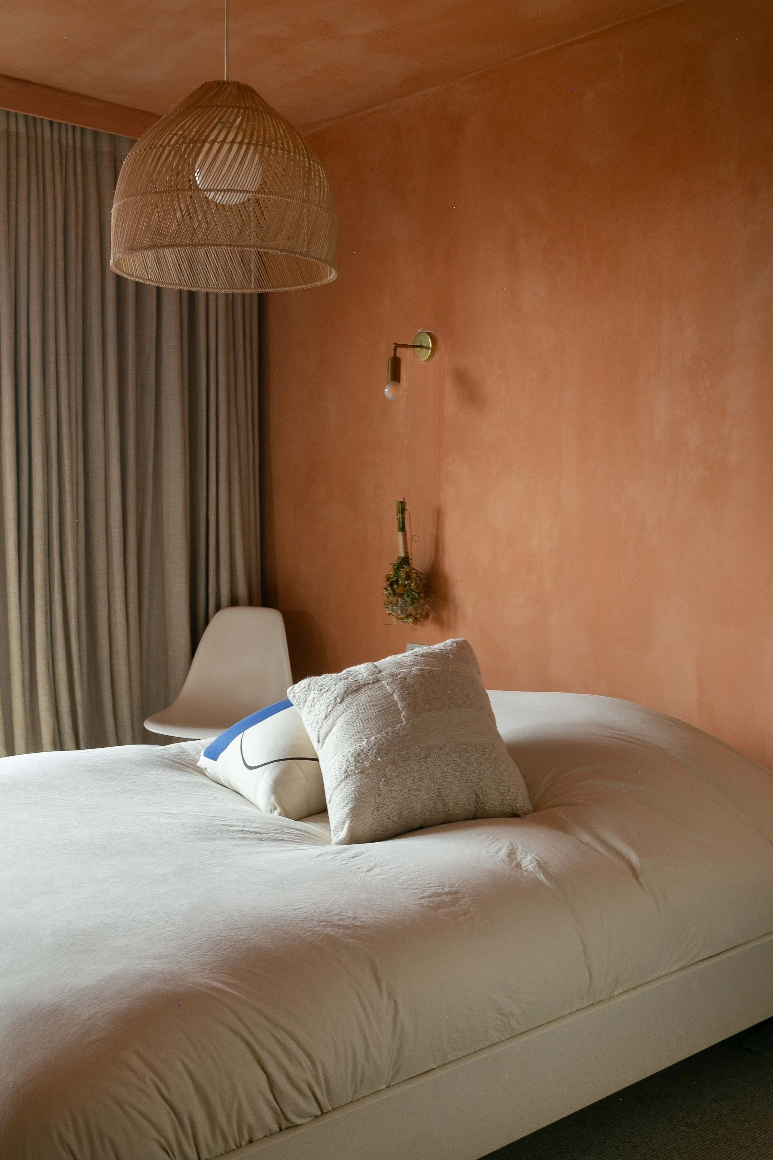 A bedroom with a white bed, beige pillows, a desk chair, a woven pendant light, and a pinkish-orange wall with a wall sconce and dried flowers.