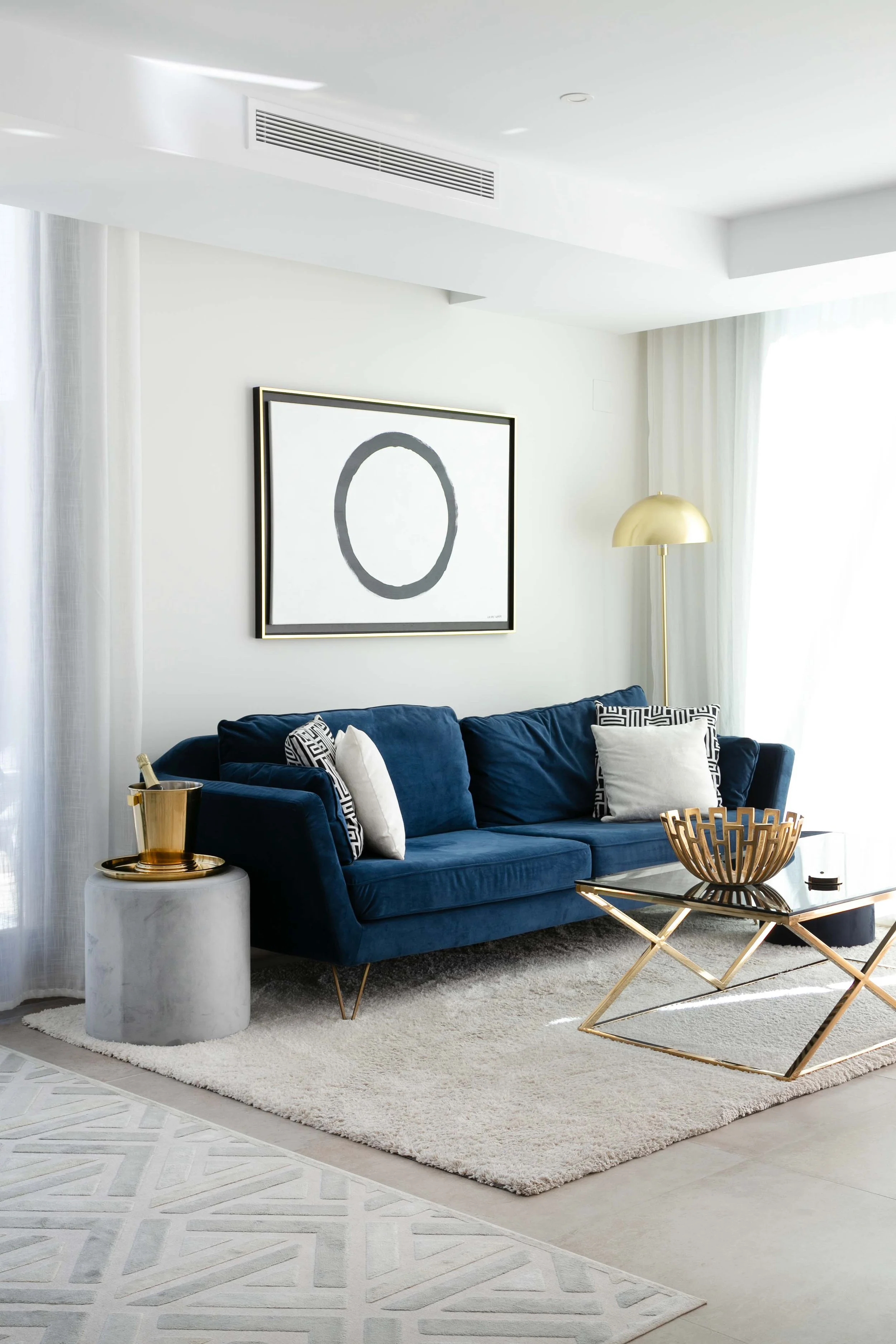 Modern living room with a royal blue velvet sofa, white and geometric patterned pillows, a gold and black coffee table, a white textured area rug, a large gold floor lamp, and minimalist wall art with a circular design, white curtains, and a light-co