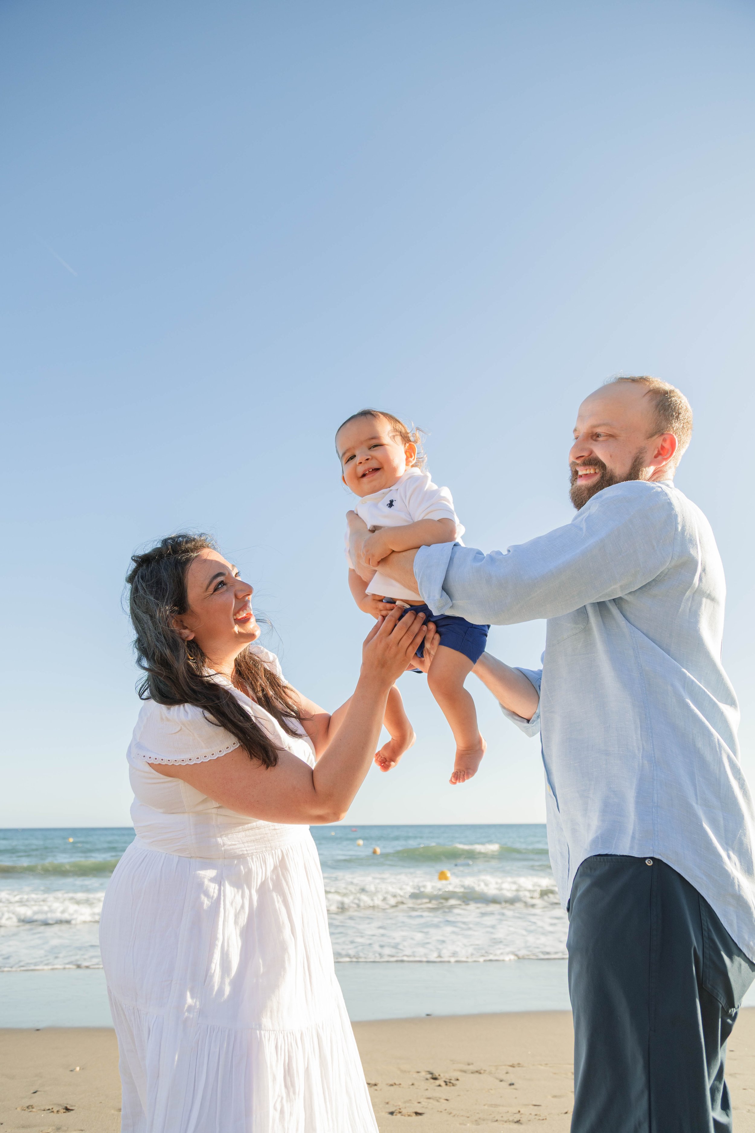 A family of three enjoying a day at the beach, with a mother holding a smiling baby, and a father reaching out to him, all under a clear blue sky.