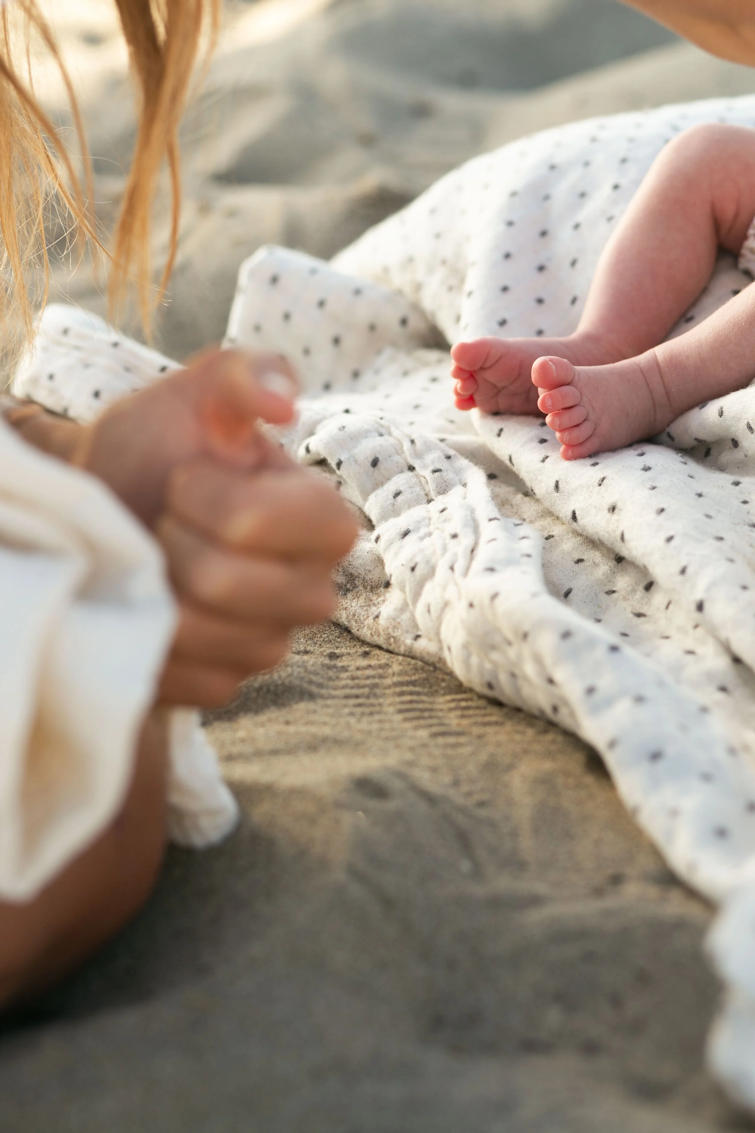 Close-up of a newborn baby's feet, lying on a white blanket with black polka dots, with a woman (likely the mother) reaching out with her hand in the foreground, on a beach.