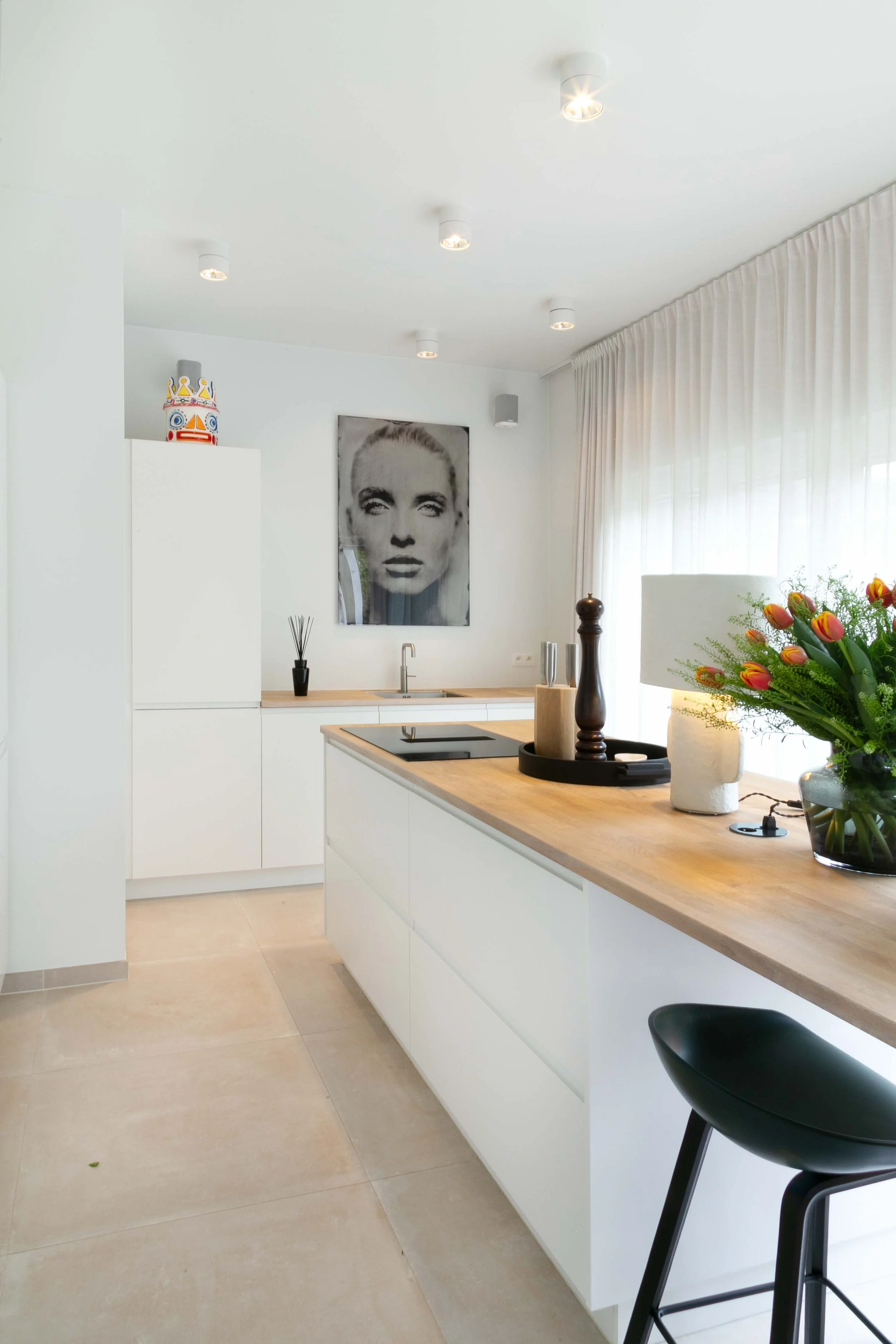 Modern minimalist kitchen with white cabinets, a wooden countertop, and a black stool. Artwork of Marilyn Monroe on the wall, and a bouquet of red and yellow tulips on the counter. Bright natural light filters through sheer white curtains.