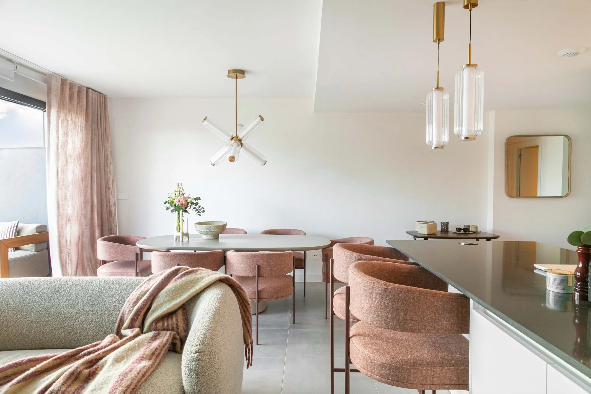 Modern dining area with pink upholstered chairs surrounding a white oval table. A floral centerpiece and decorative bowls sit on the table. Pendant lights hang from the ceiling, and a mirror is on the wall. A large window with pink curtains allows na