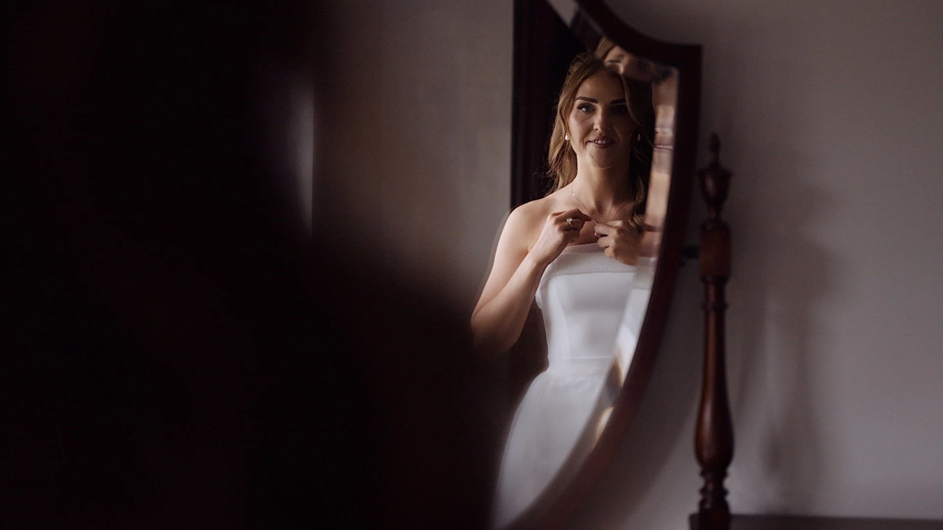Wedding film showing a woman with red hair looking at herself in a mirror, adjusting her necklace in a room with neutral-colored walls.