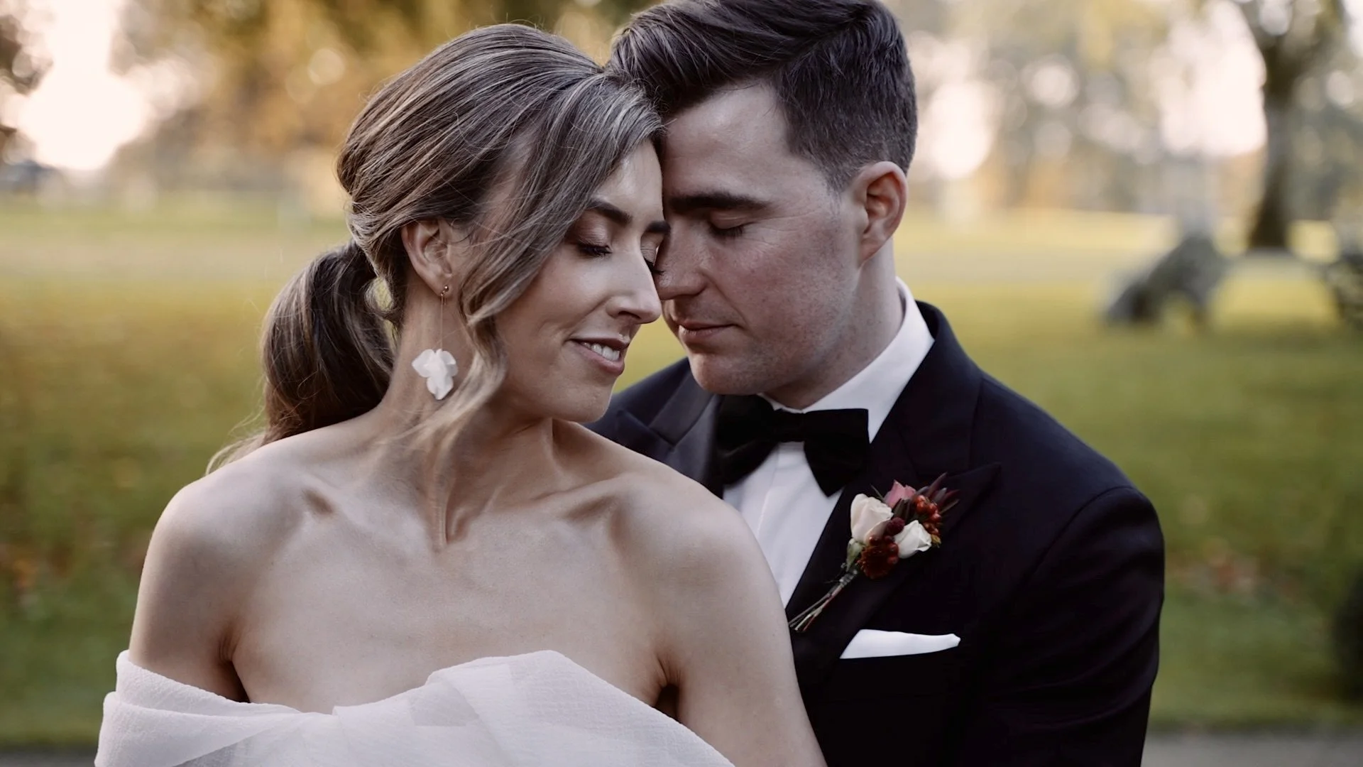 A bride and groom share an intimate moment outdoors, their foreheads touching and eyes closed, during a wedding photoshoot in a park with autumn foliage in the background.