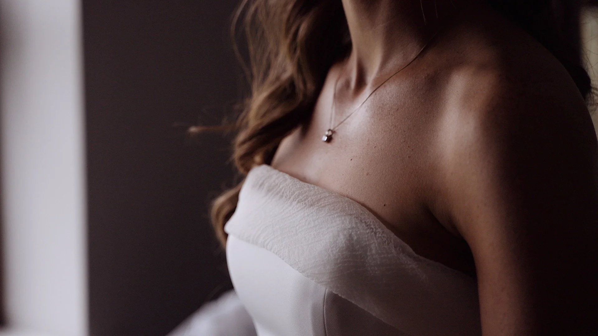 Wedding film close-up of a woman's bare shoulder, neck, and part of her face, with brown wavy hair and a delicate necklace, wearing a white off-shoulder top.