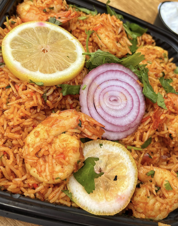 Close-up of shrimp fried rice garnished with lemon slices, a slice of red onion, and cilantro in a black takeout container.
