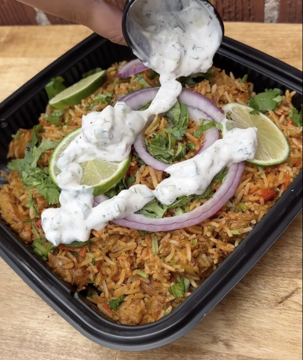 A takeout container filled with Mexican-style rice topped with sliced red onions, cilantro, and lime wedges, with a drizzle of creamy white sauce being poured on top.