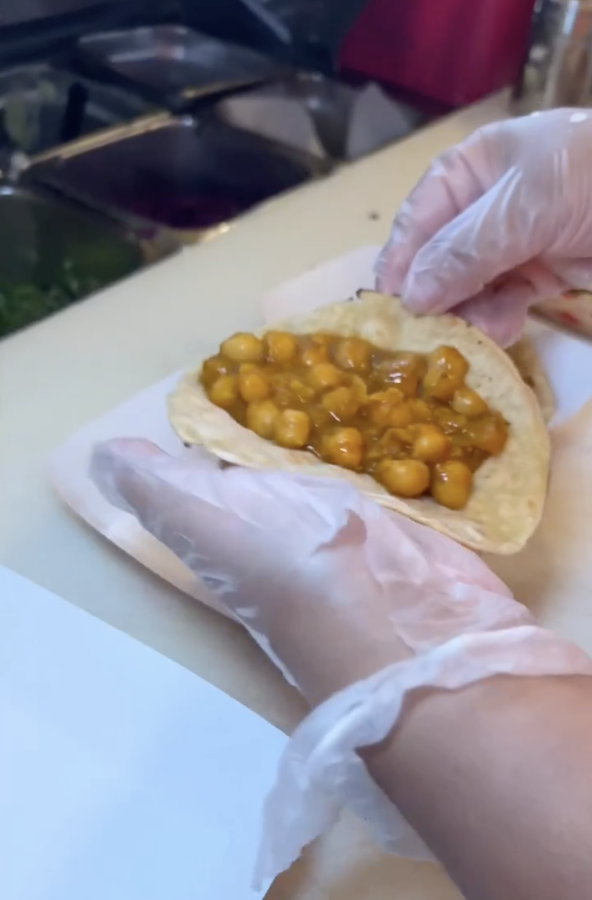 Person wearing gloves preparing a chickpea sandwich or wrap with chickpeas and sauce.