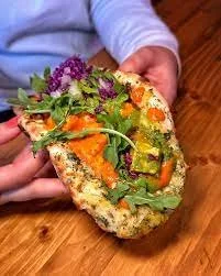 Person holding a half-eaten vegetable and hummus stuffed pita sandwich with lettuce, cherry tomatoes, purple cabbage, and herbs on a wooden table.