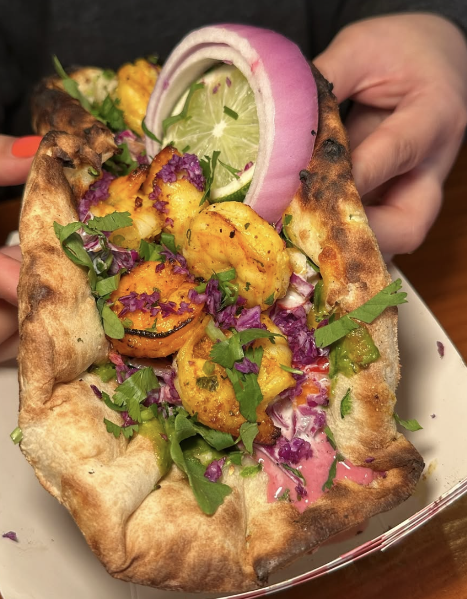 Close-up of a hand holding a flatbread topped with grilled shrimp, purple cabbage, chopped herbs, and slices of lime, garnished with onion and greens.