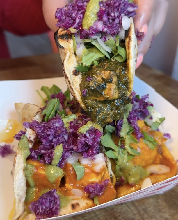 Close-up of a person holding a taco filled with spinach, purple shredded cabbage, and a lemon wedge, with more tacos on a white plate topped with purple cabbage, lettuce, green sauce, and orange sauce.