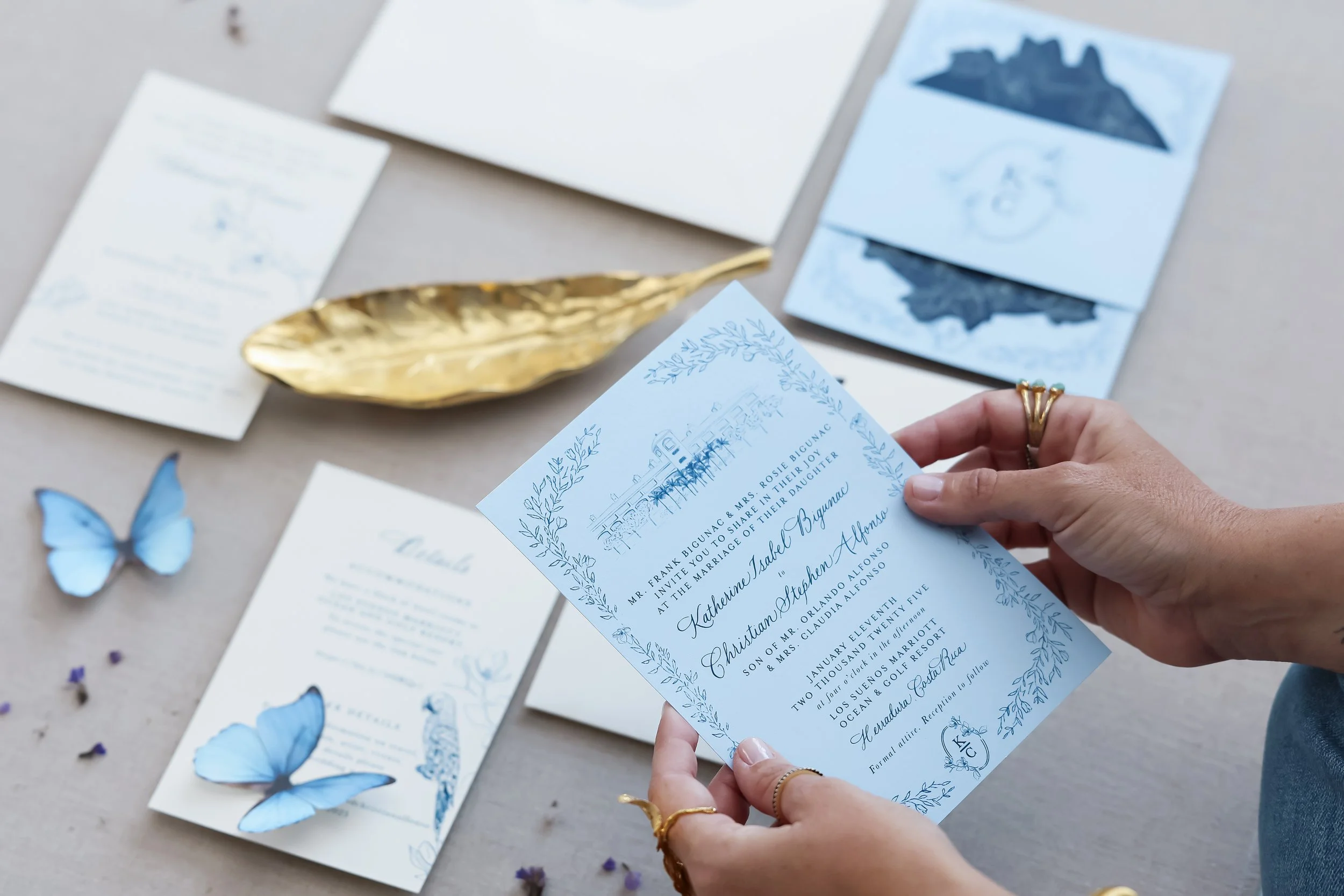 A person holding a luxury blue wedding invitation with floral and venue illustrations, surrounded by matching invitation cards,