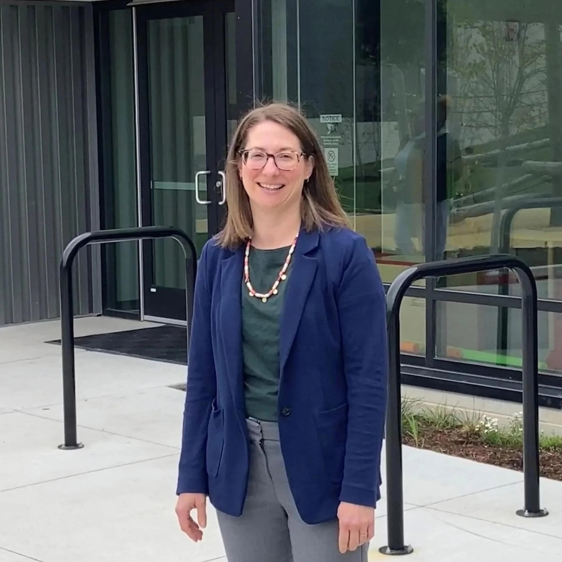 A woman with glasses, wearing a blue blazer and gray pants, standing outdoors in front of a glass building entrance with a notice sign. She is smiling and has shoulder-length brown hair.