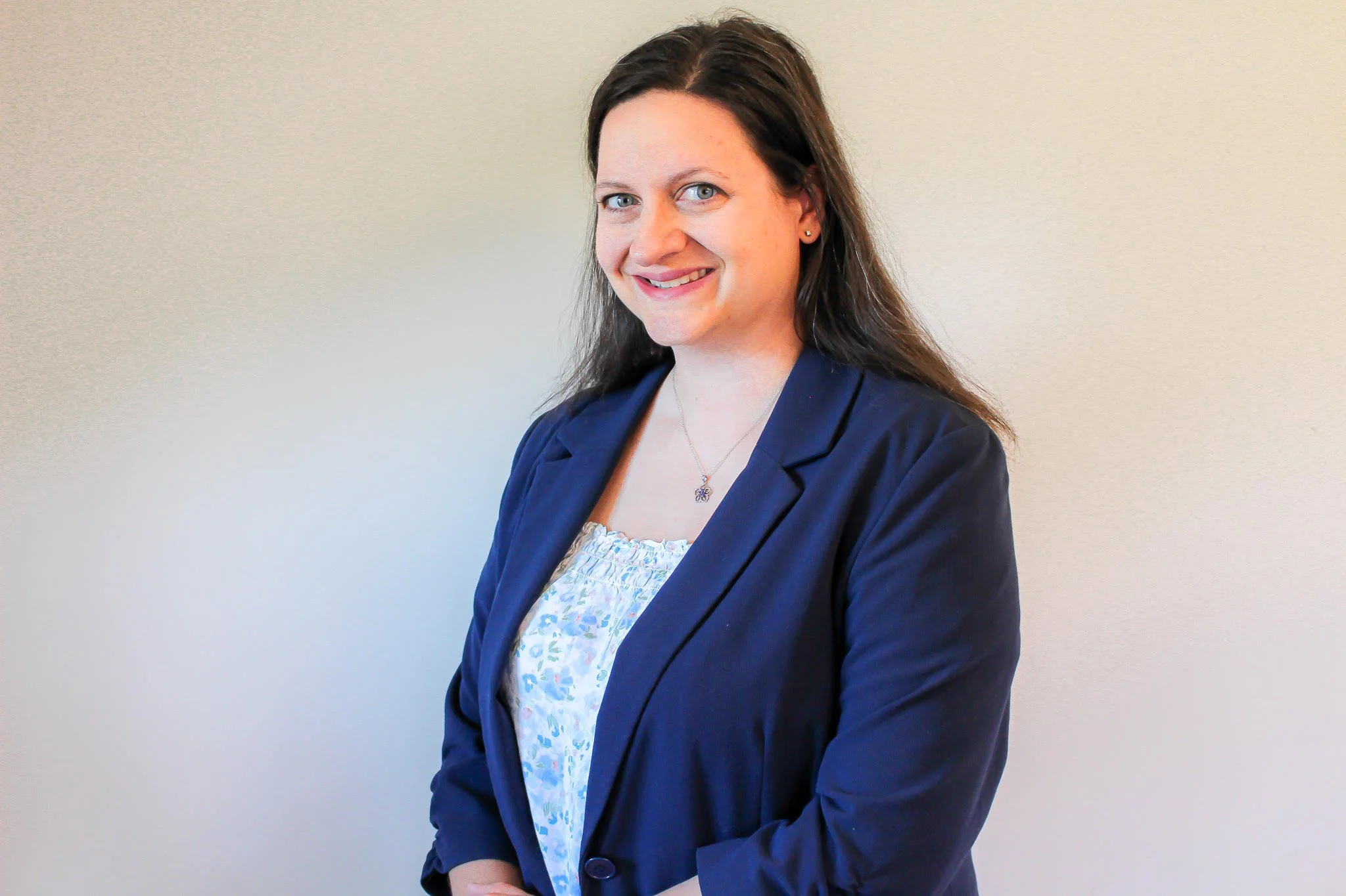 A woman with long brown hair, blue eyes, and fair skin, wearing a blue blazer and a floral blouse, standing against a plain light-colored wall and smiling at the camera.