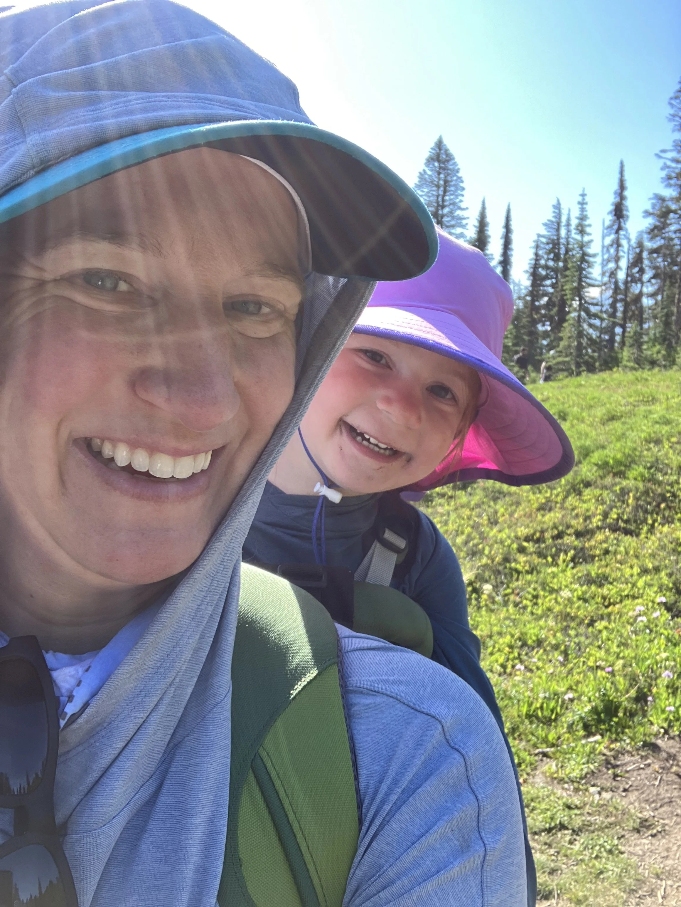 Close-up of two smiling children wearing hats and outdoor clothing, in a green forested area with blue sky.