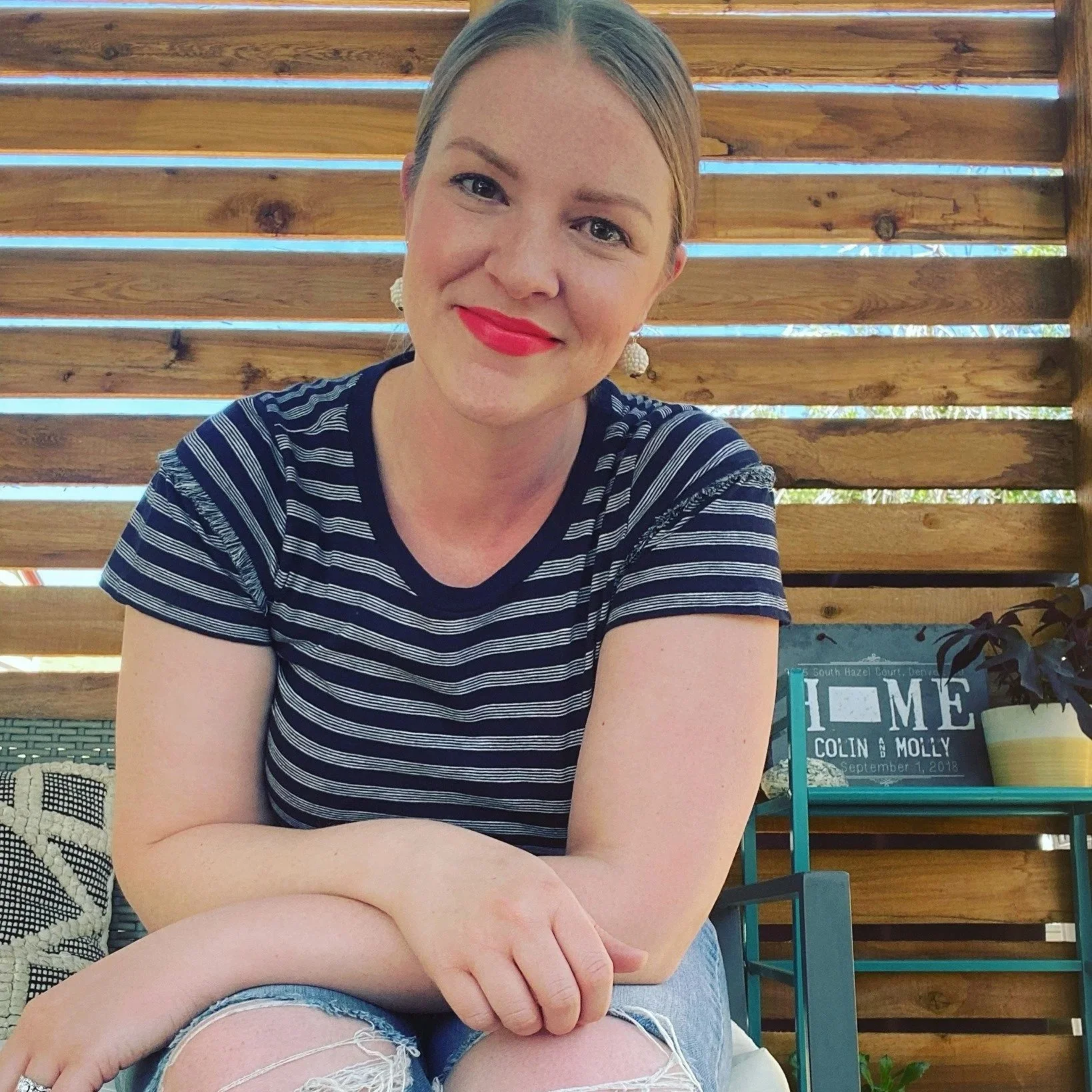 A smiling woman with short light brown hair, red lipstick, and pearl earrings, wearing a blue and white striped shirt and ripped denim shorts, sitting outdoors in front of a wooden fence.