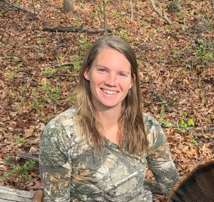 Woman with long hair wearing camouflage clothing, smiling, outdoors in a wooded area with brown leaves and small green plants.