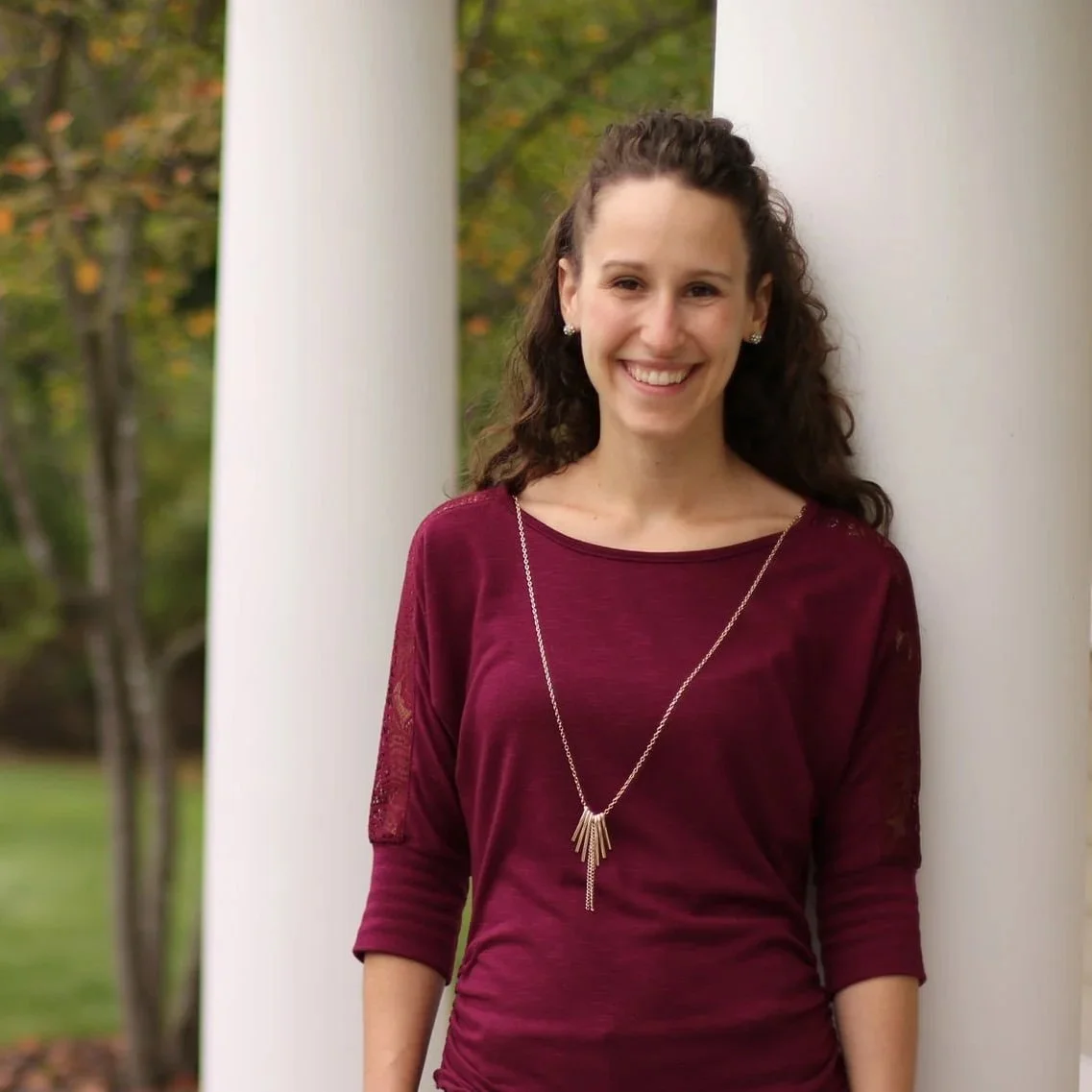 A young woman with curly brown hair smiling, wearing a maroon top and a long silver necklace, standing outdoors next to white columns with trees and autumn leaves in the background.