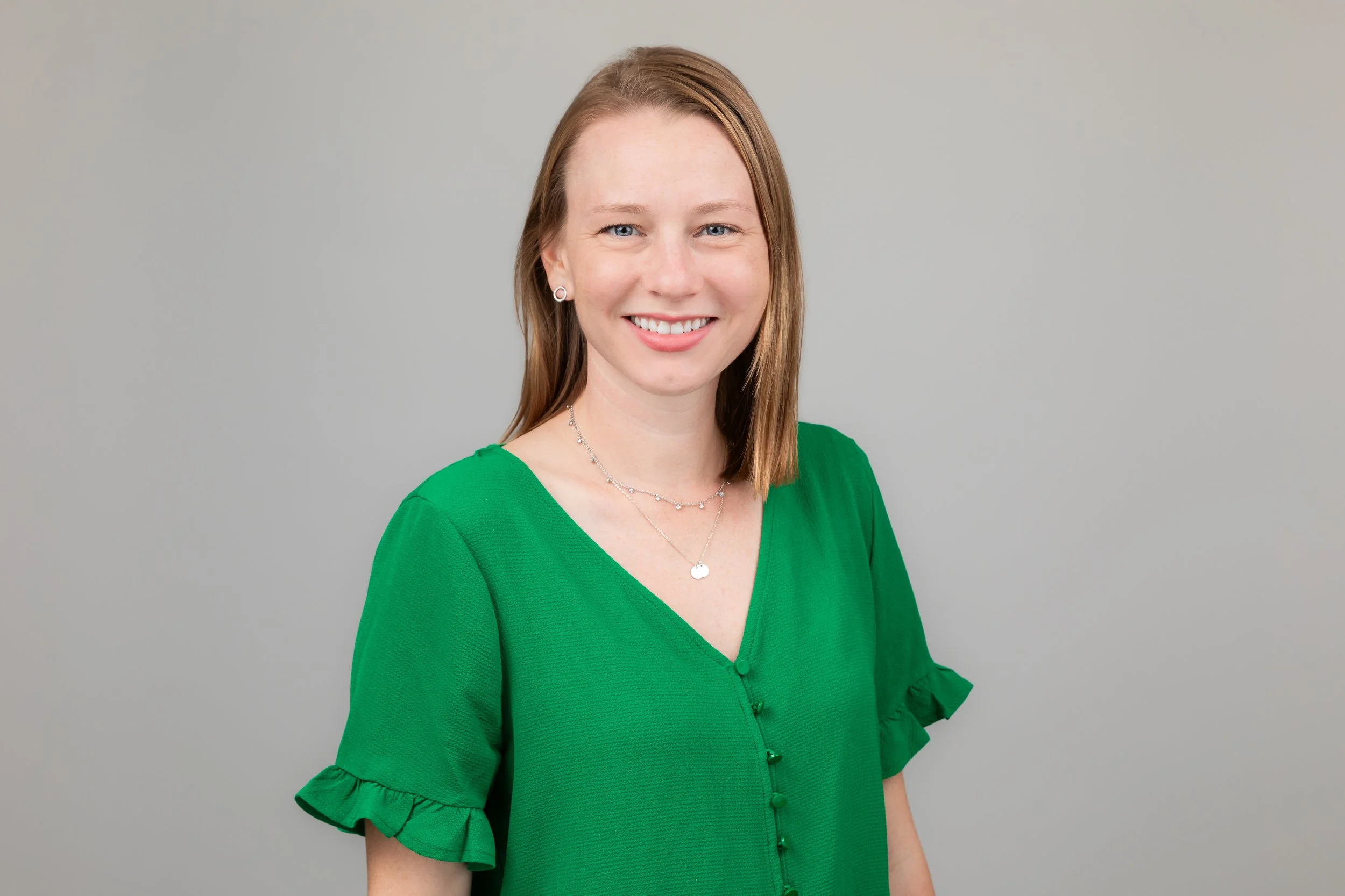 A young woman with shoulder-length reddish hair, blue eyes, and fair skin, smiling, wearing a green blouse with ruffled sleeves, and jewelry including earrings and layered necklaces, standing against a plain light gray background.