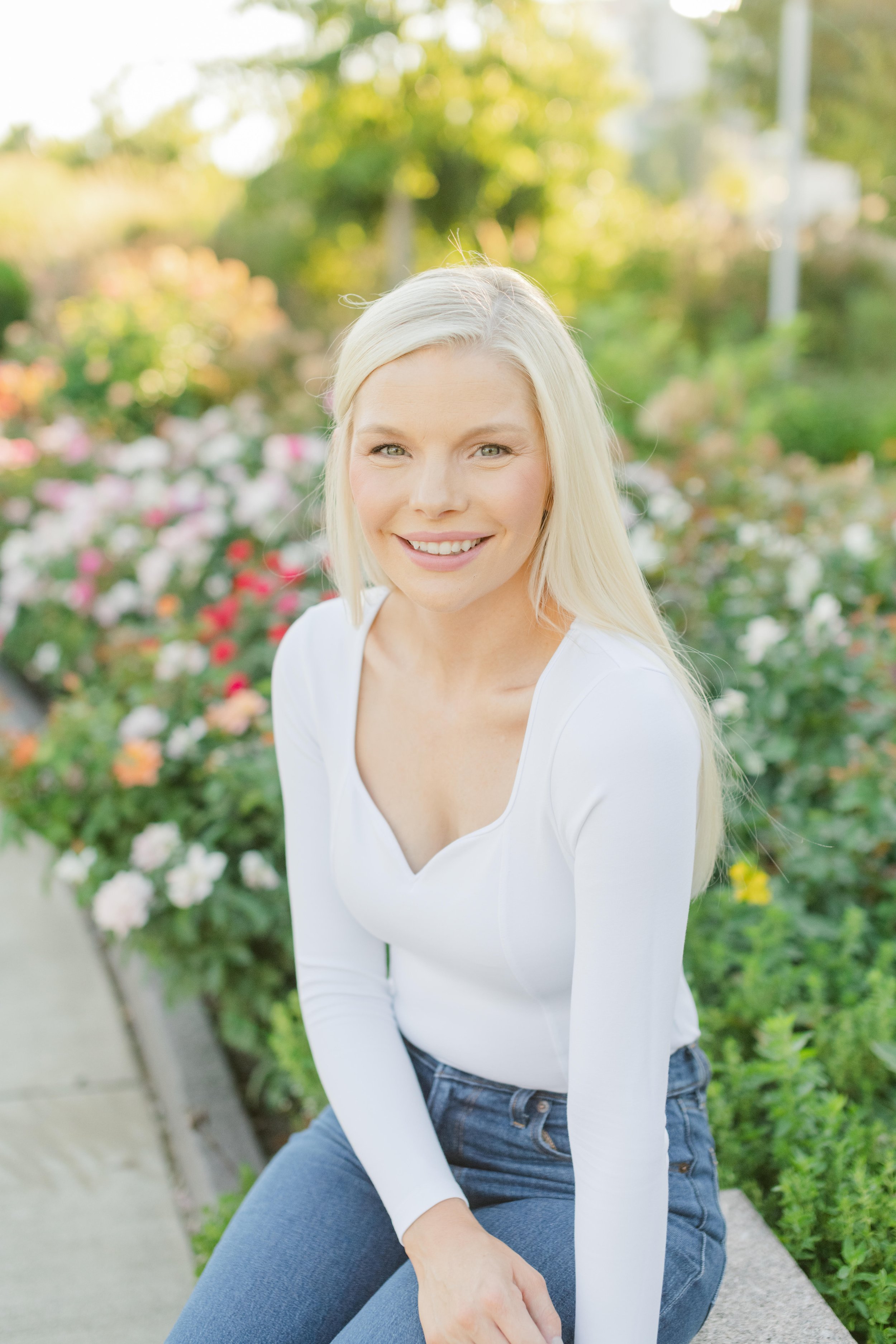 A woman with long blonde hair smiling while sitting on a concrete bench outdoors with colorful flowers and greenery in the background.