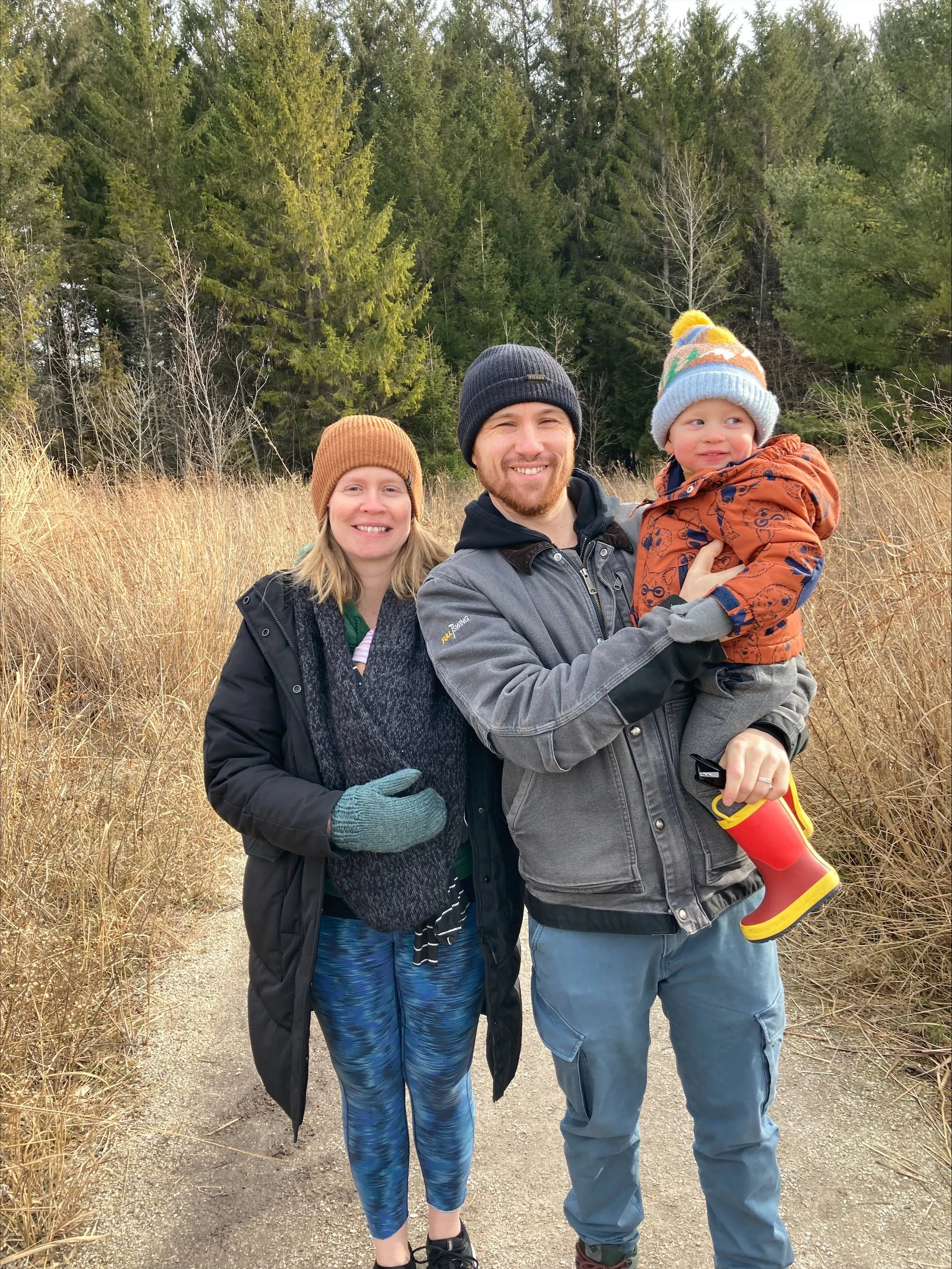 A family outdoors in a natural setting with tall grasses and pine trees. A woman, man, and young child are smiling, with the man holding the child. The woman has blonde hair and is wearing a brown knit hat, black jacket, and blue leggings. The man has a beard, is wearing a gray jacket and dark beanie. The child is dressed in an orange jacket, colorful knit hat, and gray pants, holding a yellow and red toy.