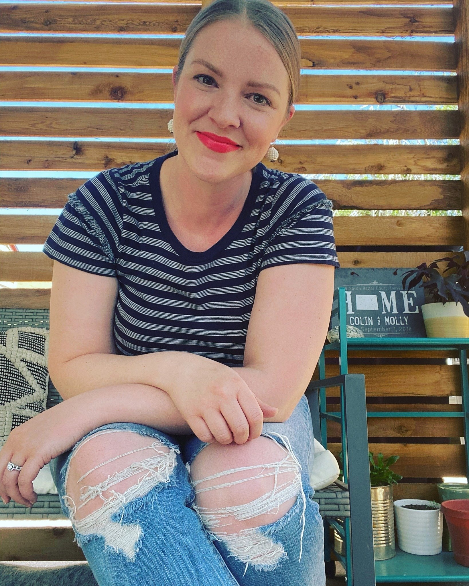 A woman with blonde hair styled with a side part, wearing red lipstick, a navy blue and white striped t-shirt, and ripped denim shorts, sitting outdoors in front of a wooden slat fence with potted plants and outdoor furniture visible nearby. She is smiling and looking at the camera.