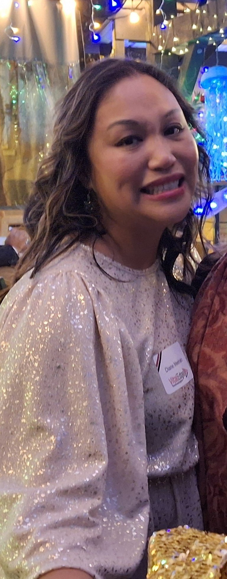 A woman with wavy brown hair smiling at a festive gathering, wearing a sparkly light-colored top with shiny glitter details, and standing in a setting decorated with colorful string lights and holiday decorations.